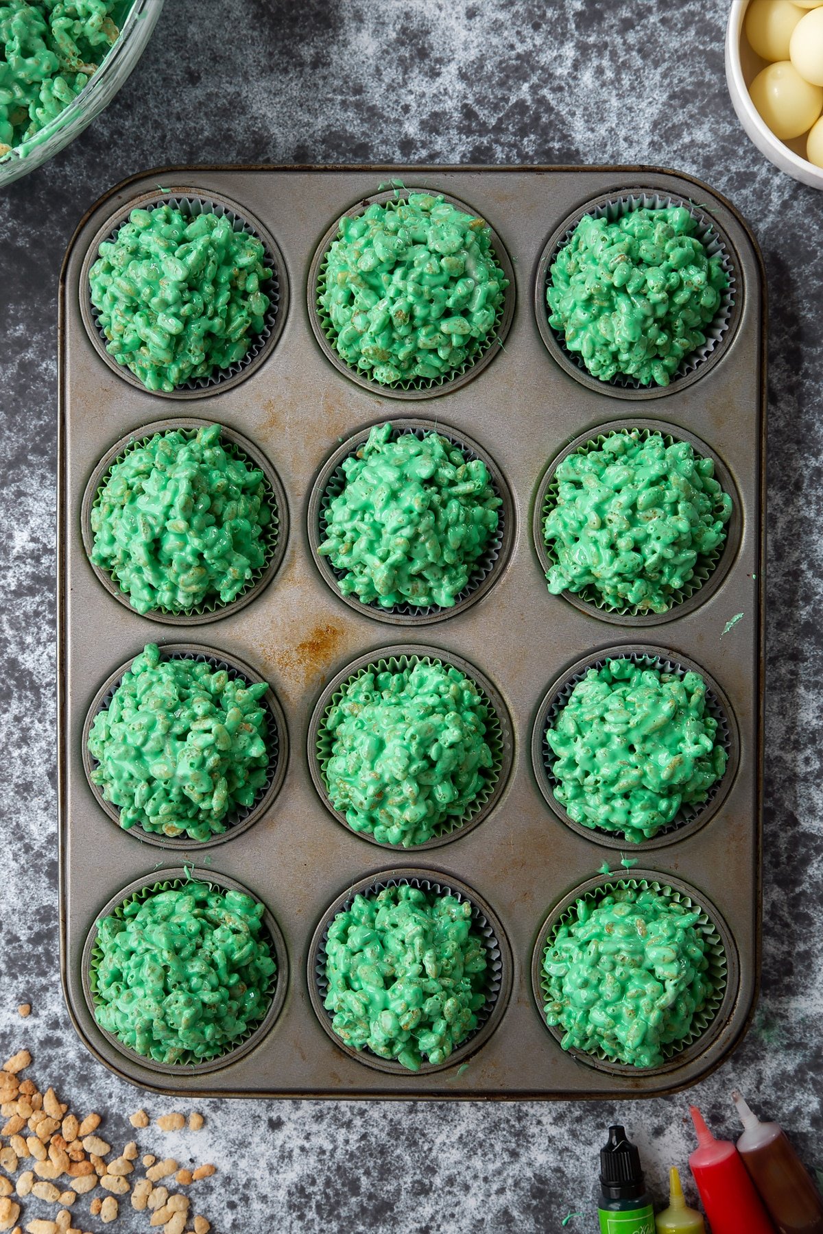 12-hole muffin tray lined with cupcake cases filled with green rice crispy treats. Ingredients to make Halloween crispy cakes surround the tray.