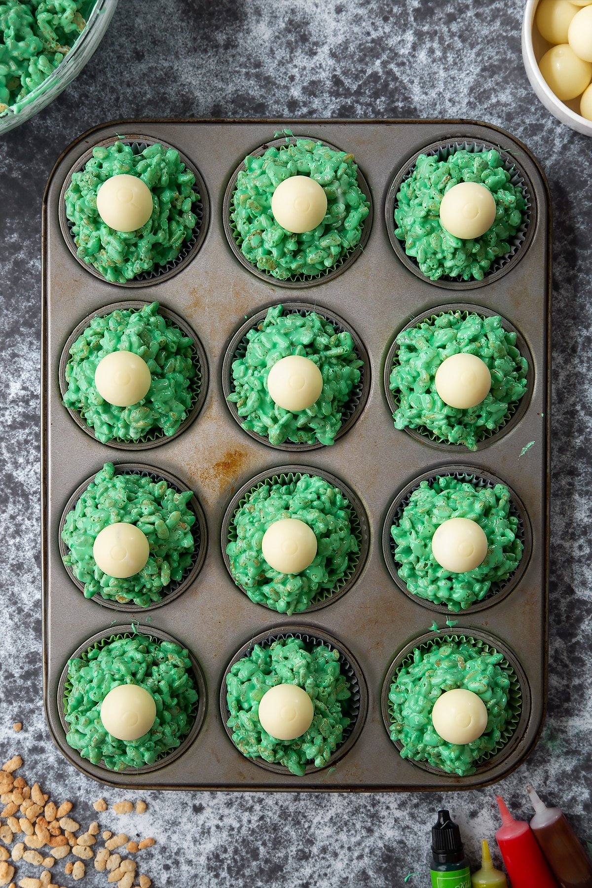 12-hole muffin tray lined with cupcake cases filled with green rice crispy treats topped with white chocolate spheres. Ingredients to make Halloween crispy cakes surround the tray.