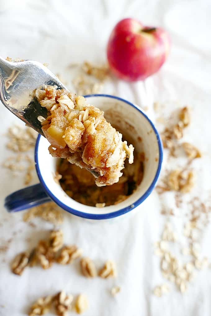 An apple crumble in a mug with an oaty topping. A fork takes a helping of the mug dessert.