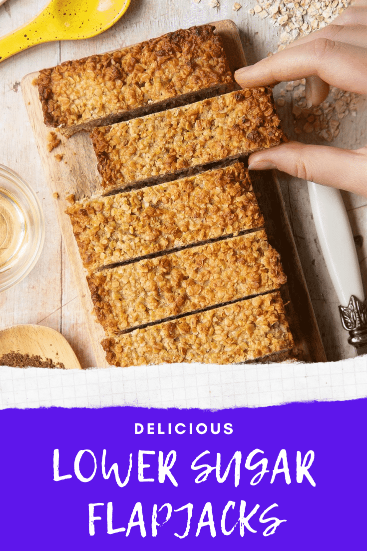 Overhead shot of a hand holding a lower sugar flapjack served on a wooden plate with the delicious low sugar graphic at the bottom. 