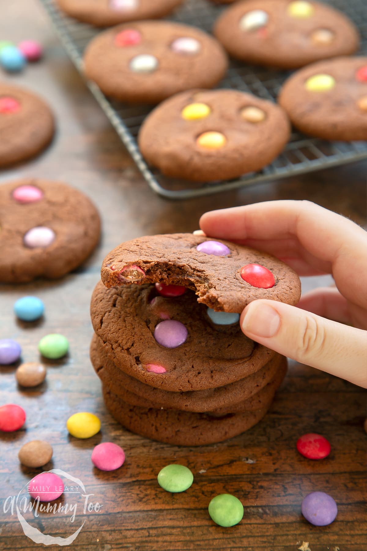 Smartie cookies stacked up on a dark wooden surface. The top cookie has been bitten and a hand reaches to take it. 