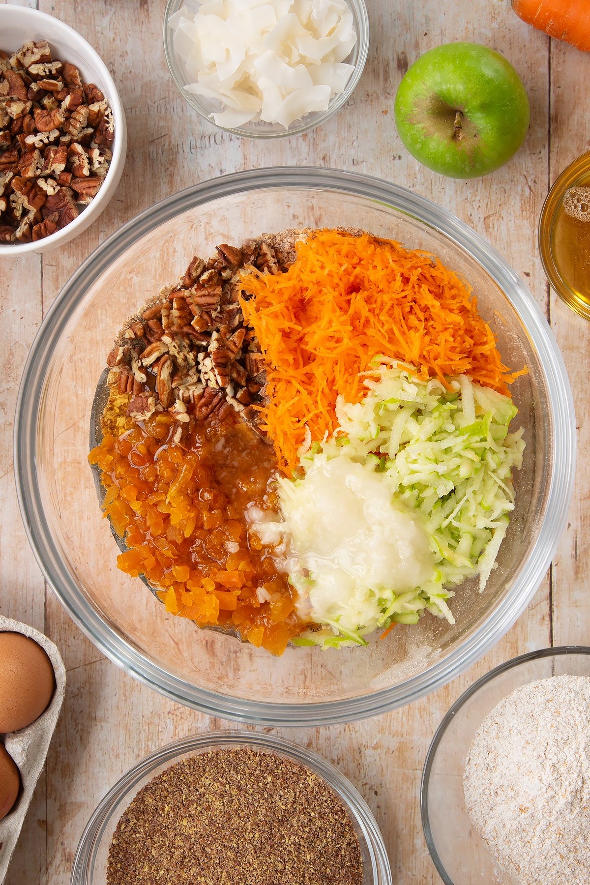 Overhead shot of dry ingredients mix, dried apricots, nuts, apple, carrot, coconut oil, maple syrup, orange zest and juice in a large clear bowl