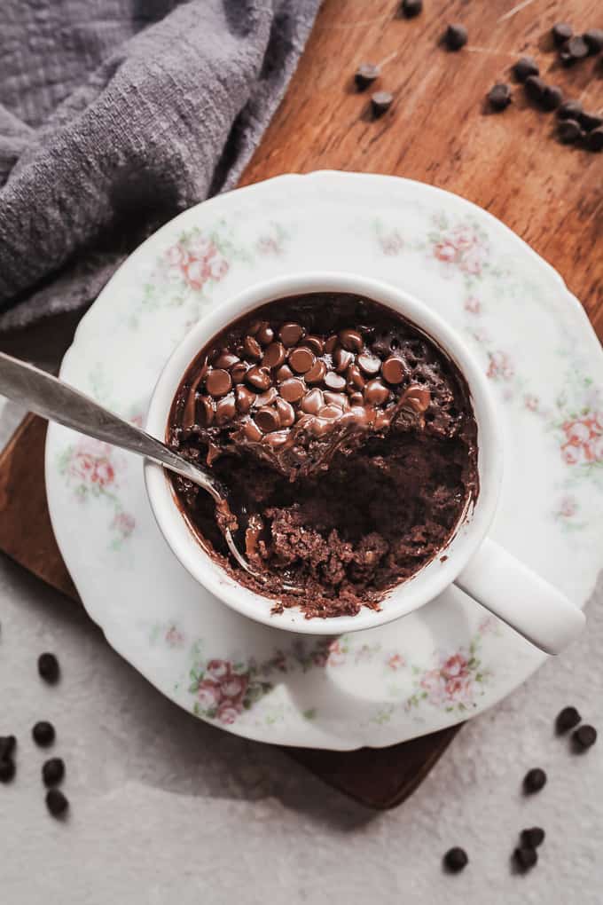 A rich, dark, vegan chocolate mug cake, topped generously with chocolate chips. A spoon delves into the cake.