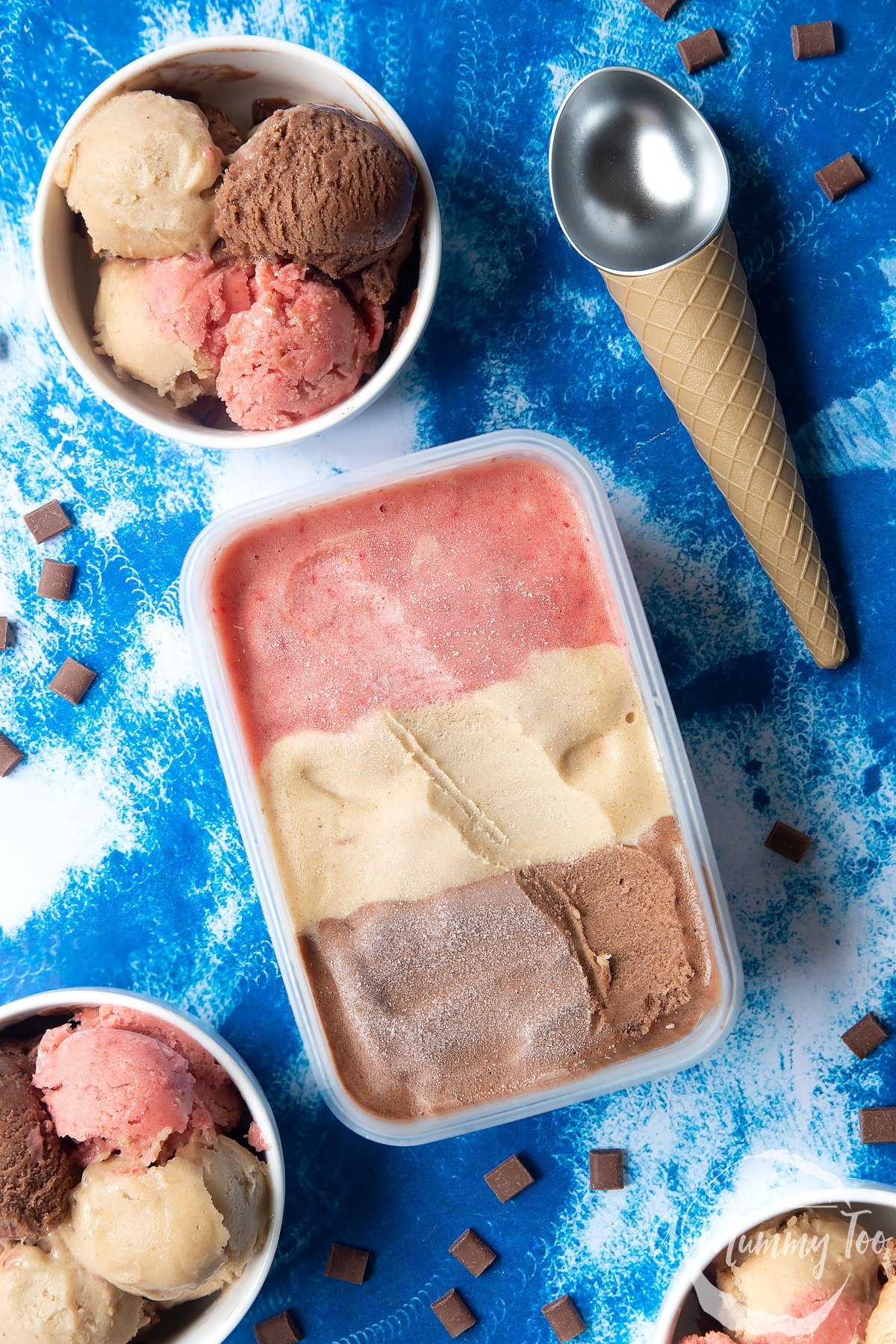 Dairy-free Neapolitan ice cream in a tub. More is served in small white bowls. An ice cream scoop and scattered chocolate chips are also shown.