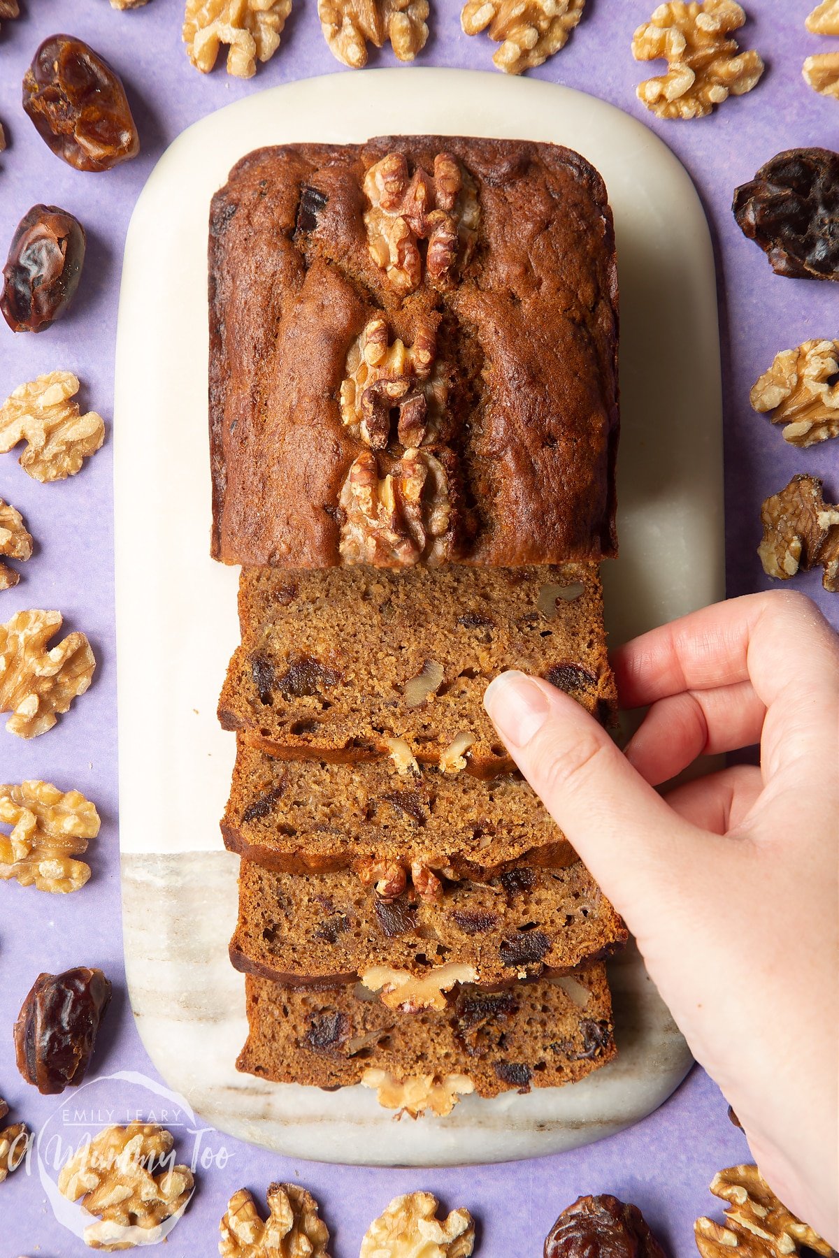 Overhead shot of  a hand holding a Vegan date and walnut cake served on a marble plate with a mummy too logo in the lower-left corner