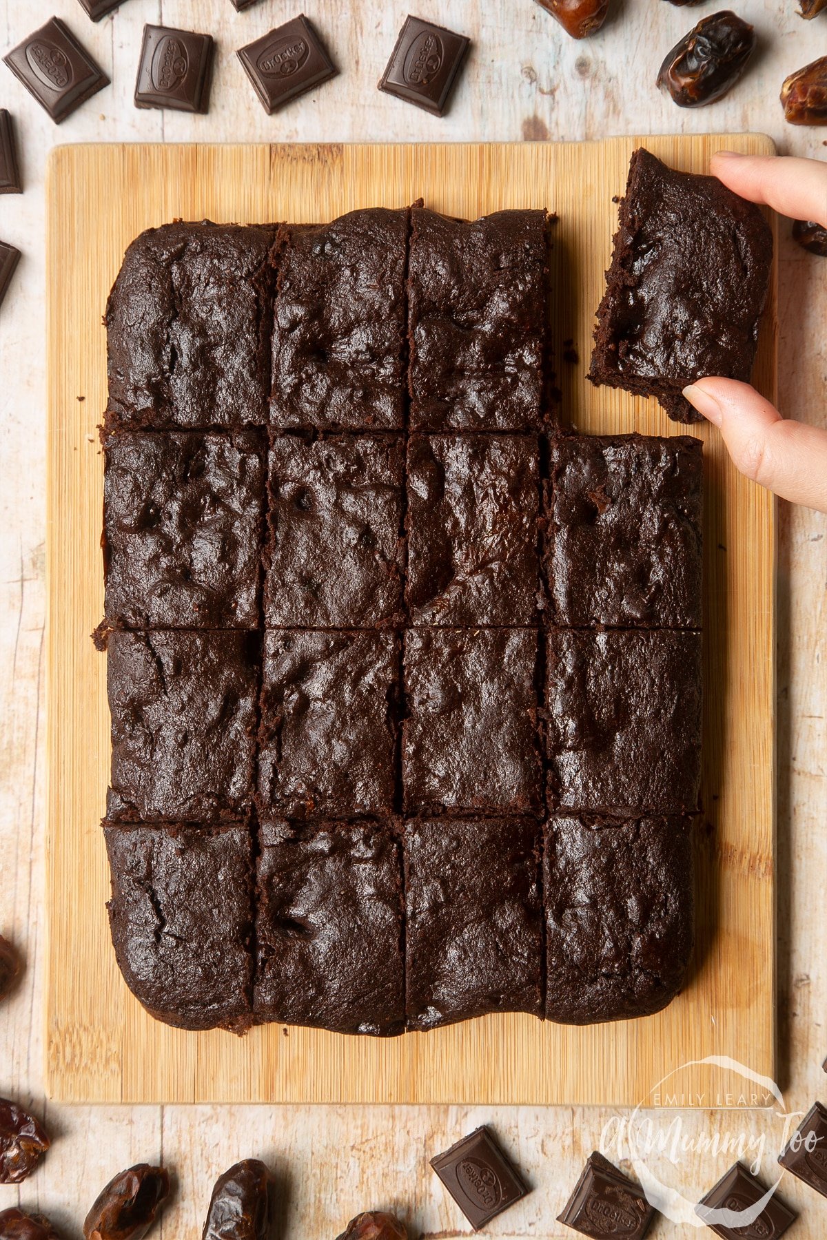 16 whole grain brownies made with dates and dark chocolate arranged on a pale wooden board. A hand reaches to take one.