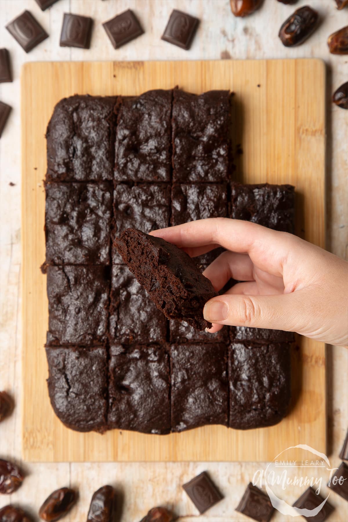Whole grain brownies made with dates and dark chocolate arranged on a pale wooden board. A hand holds one.