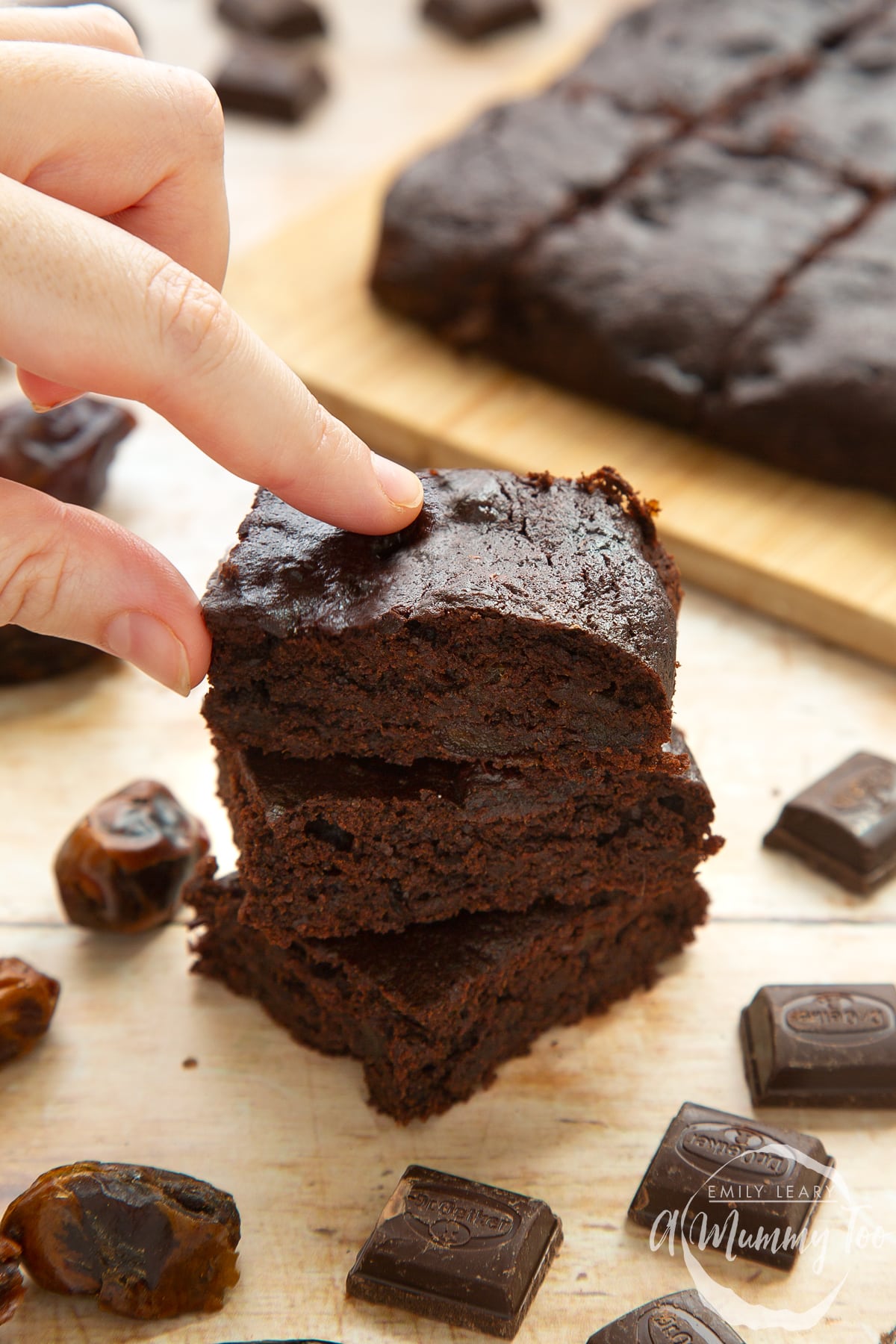 Whole grain brownies made with dates and dark chocolate, stacked up on pale wood. A hand reaches to take one.