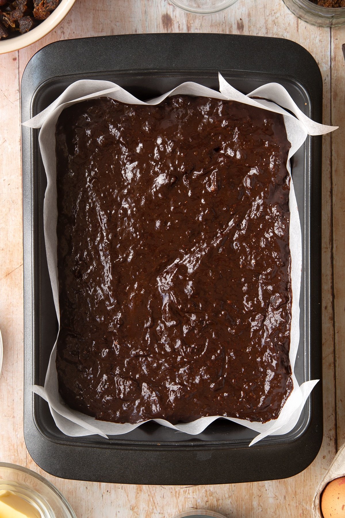 A rectangular tin lined with baking paper and filled with whole grain brownie batter. Ingredients to make whole grain brownies surround the tin.