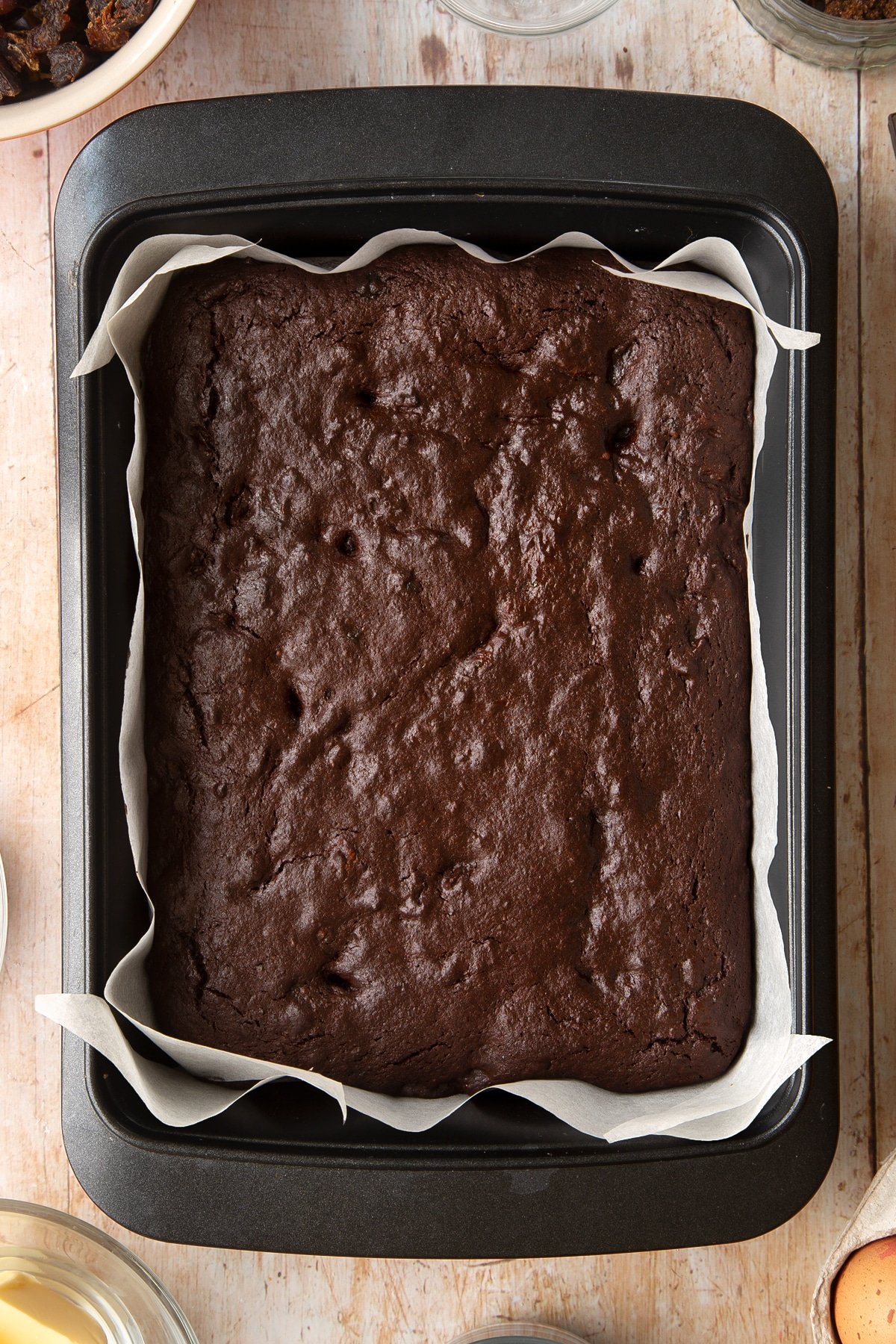 A rectangular tin lined with baking paper and containing freshly baked whole grain brownies. Ingredients to make whole grain brownies surround the tin.