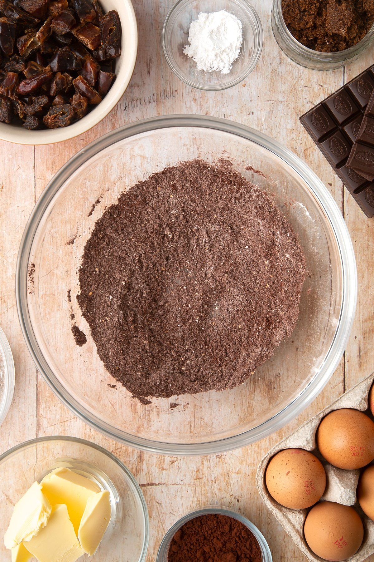 A mixing bowl containing cocoa, wholemeal flour and baking powder, whisked together. Ingredients to make whole grain brownies surround the bowl.