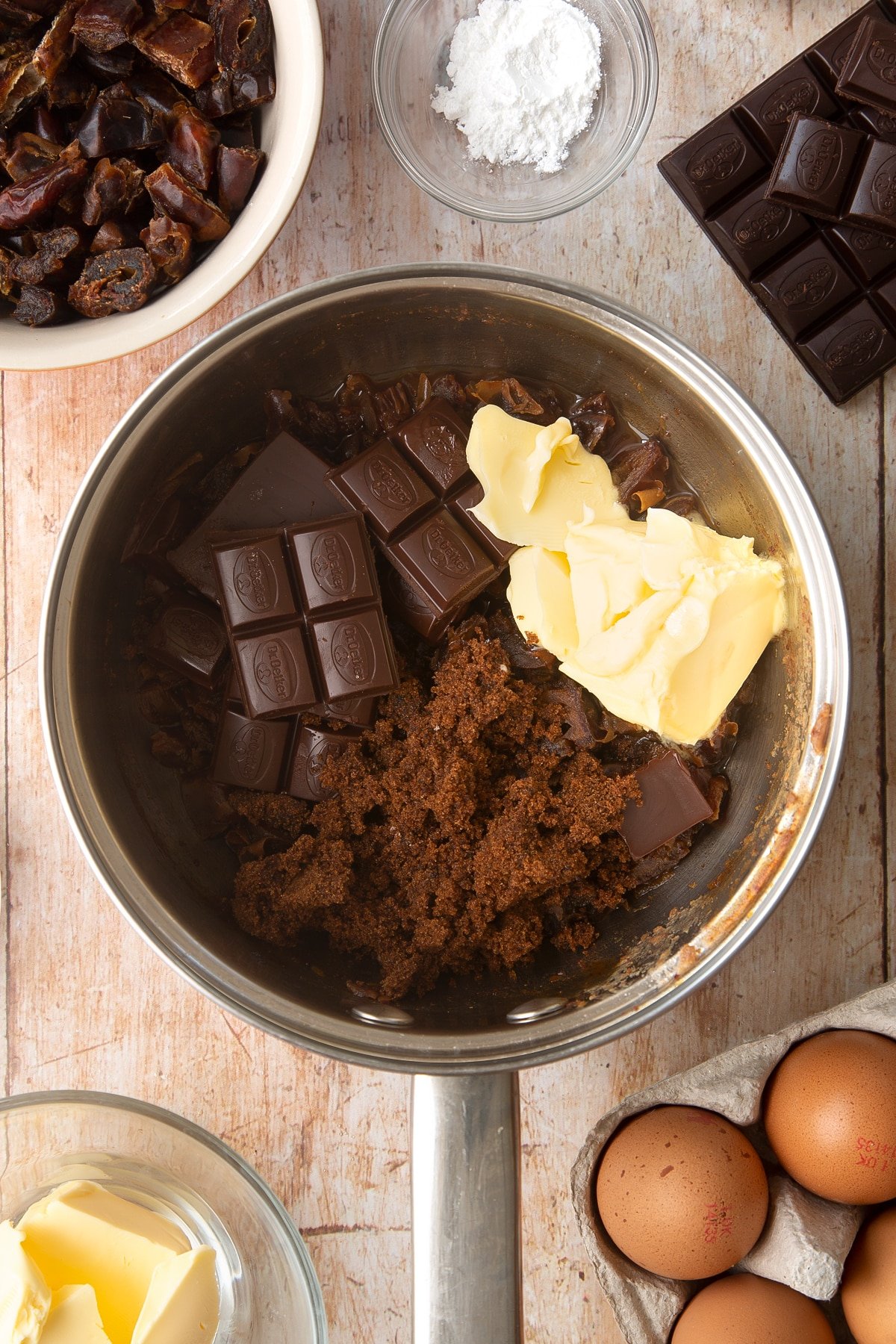 A sauce pan containing chopped dried dates simmered in water. On top sits butter, dark chocolate and sugar. Ingredients to make whole grain brownies surround the pan.