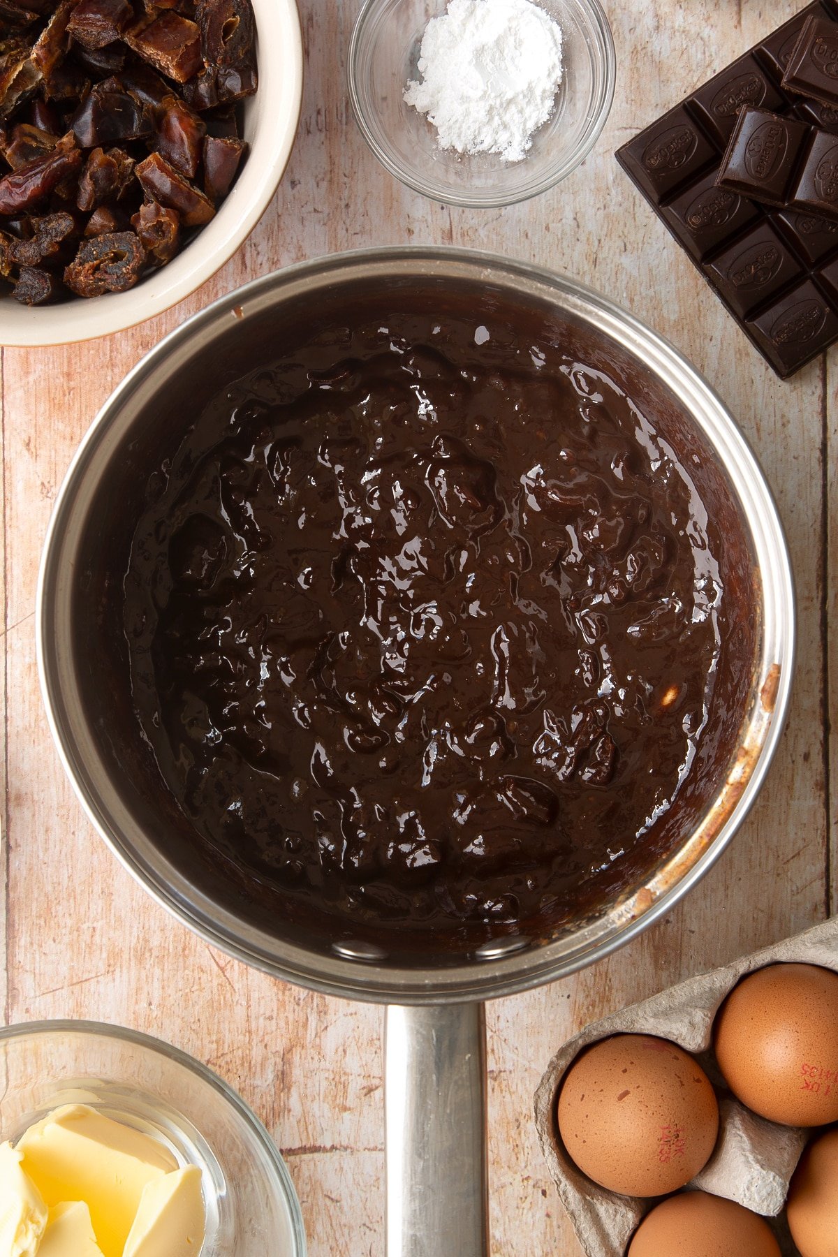 A sauce pan containing chopped dates melted together with butter, dark chocolate and sugar. Ingredients to make whole grain brownies surround the pan.