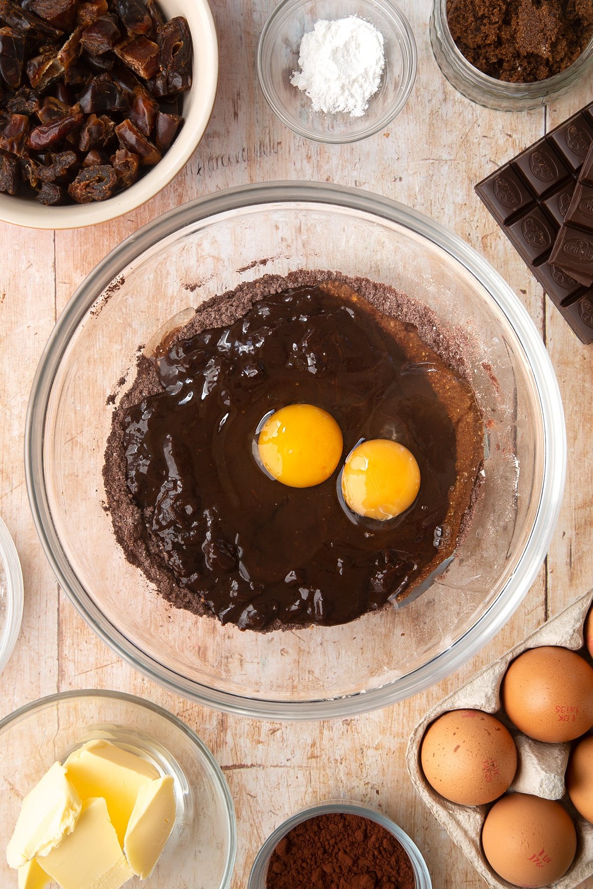 A mixing bowl containing cocoa, wholemeal flour and baking powder. The melted chocolate mixture and eggs have been added on top. Ingredients to make whole grain brownies surround the bowl.