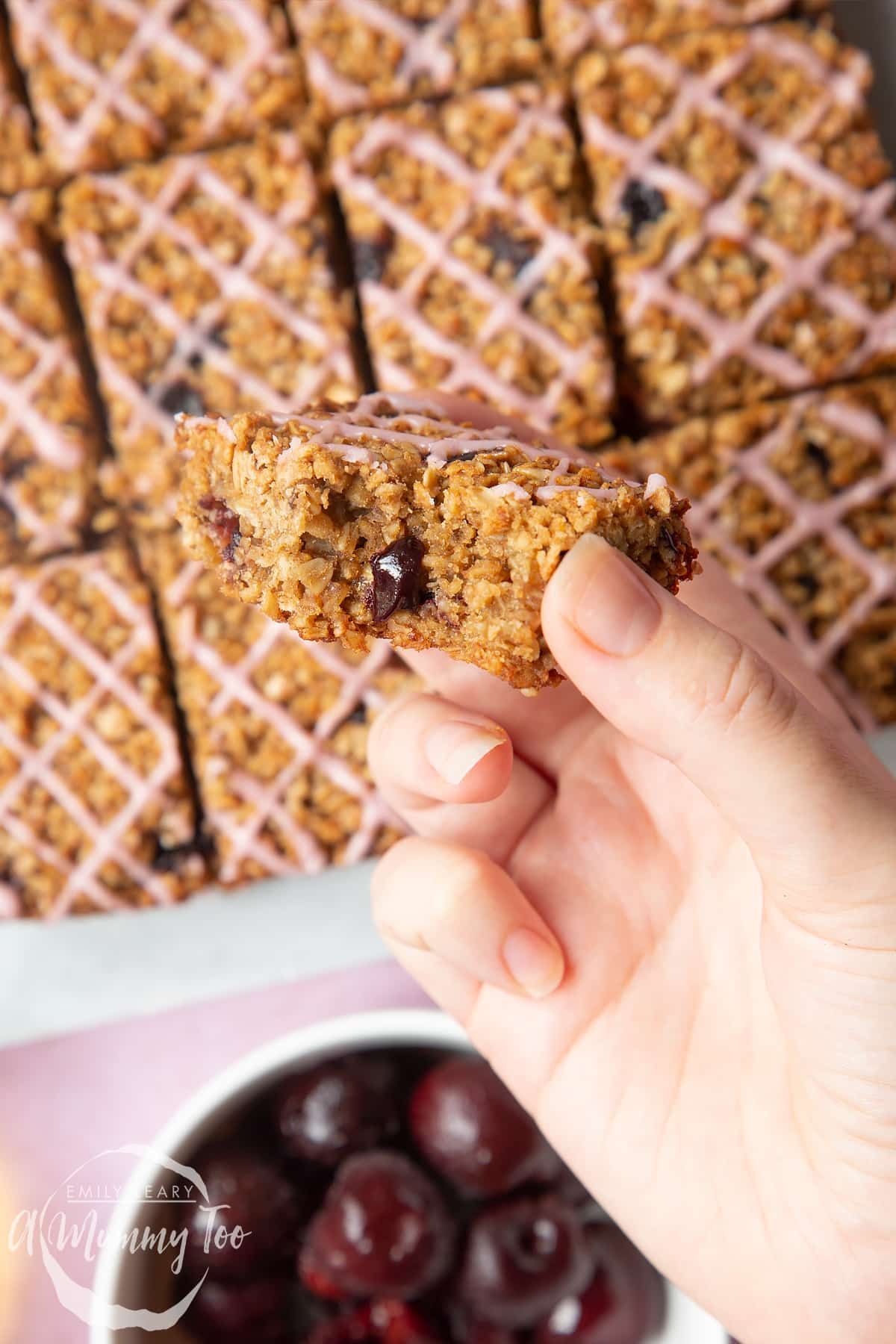 Overhead shot of a hand holding a half eaten cherry flapjack with a mummy too logo in the lower-left corner