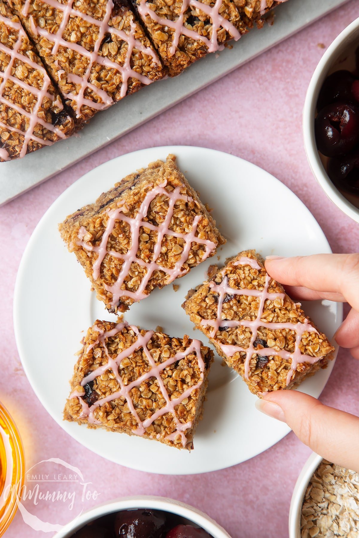 Overhead shot of a hand holding a cherry flapjack served on a white plate with a mummy too logo in the lower-left corner