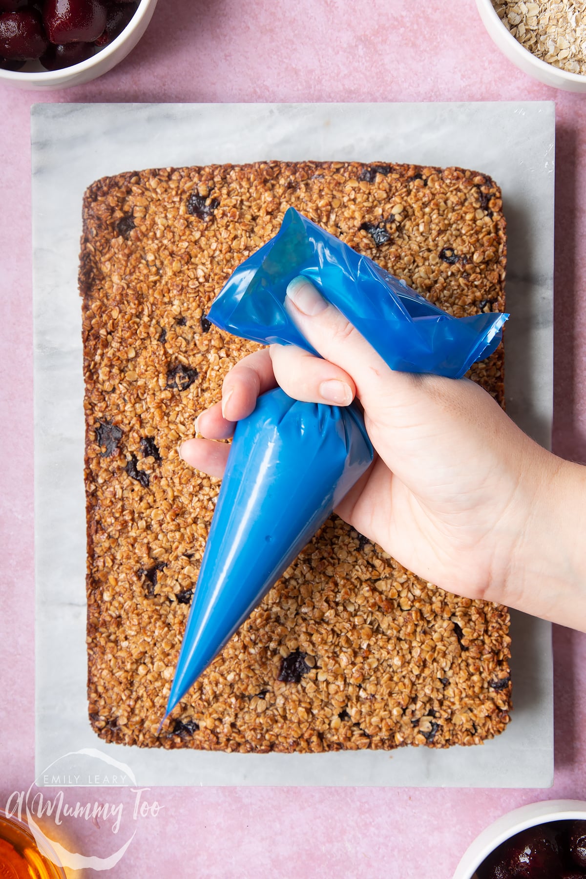 Overhead shot of a hand holding a piping bag above cherry flapjacks with a mummy too logo in the lower-left corner