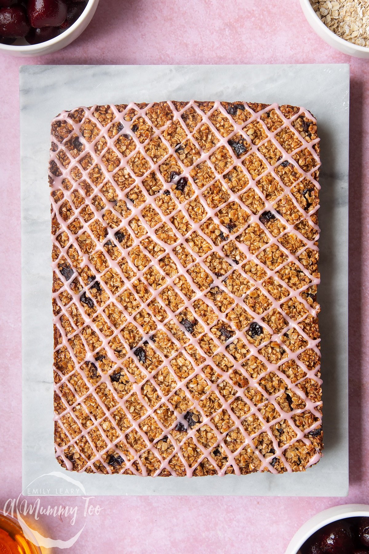 Overhead shot of cherry flapjacks topped with pink icing on a marble chopping board with a mummy too logo in the lower-left corner