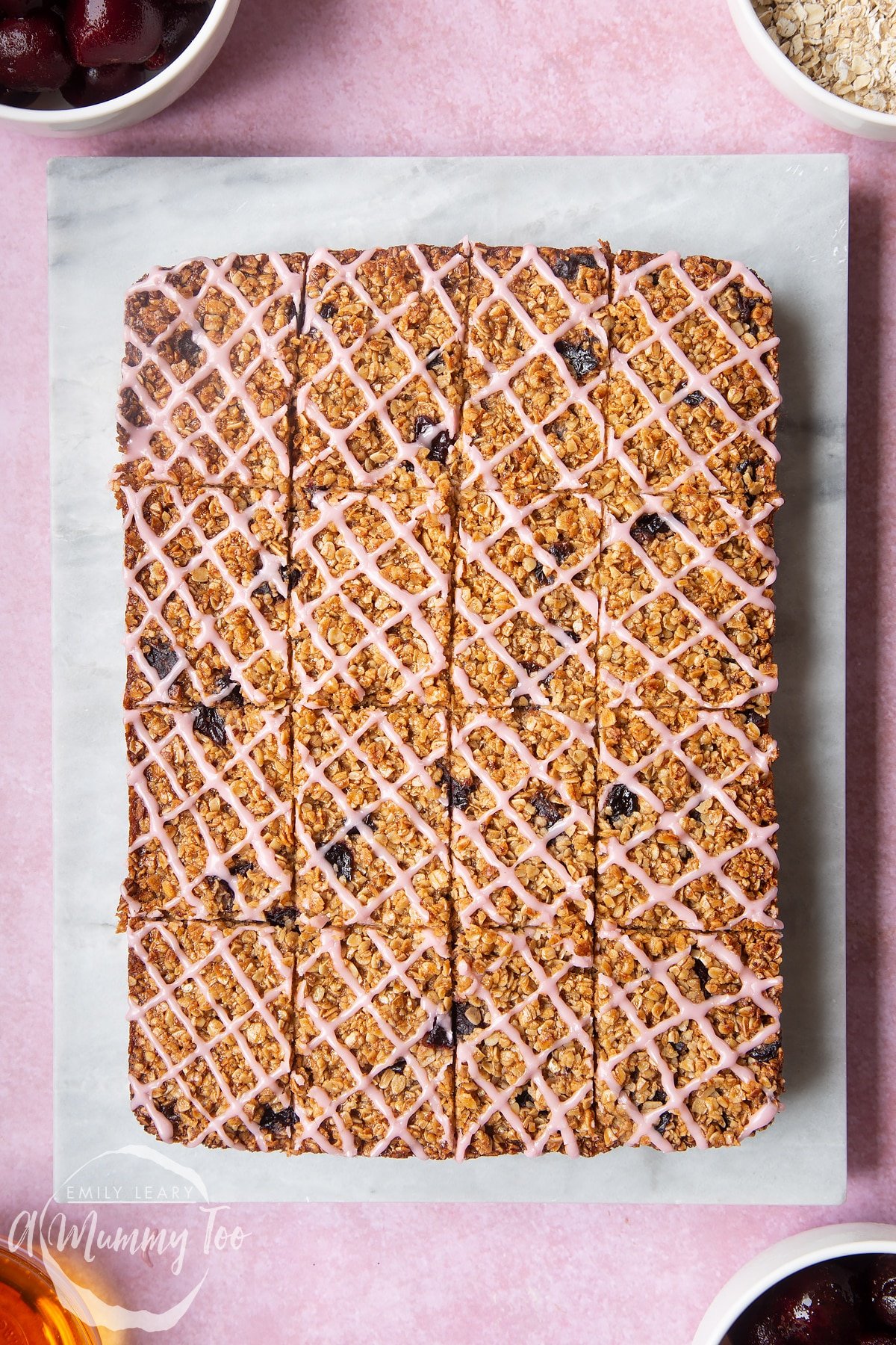 Overhead shot of cut cherry oat bars topped with pink icing on a marble chopping board with a mummy too logo in the lower-left corner