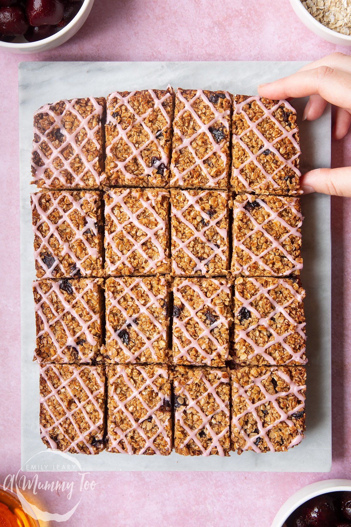 Overhead shot of a hand holding a cut cherry flapjack oat bar on a marble slab with a mummy too logo in the lower-left corner