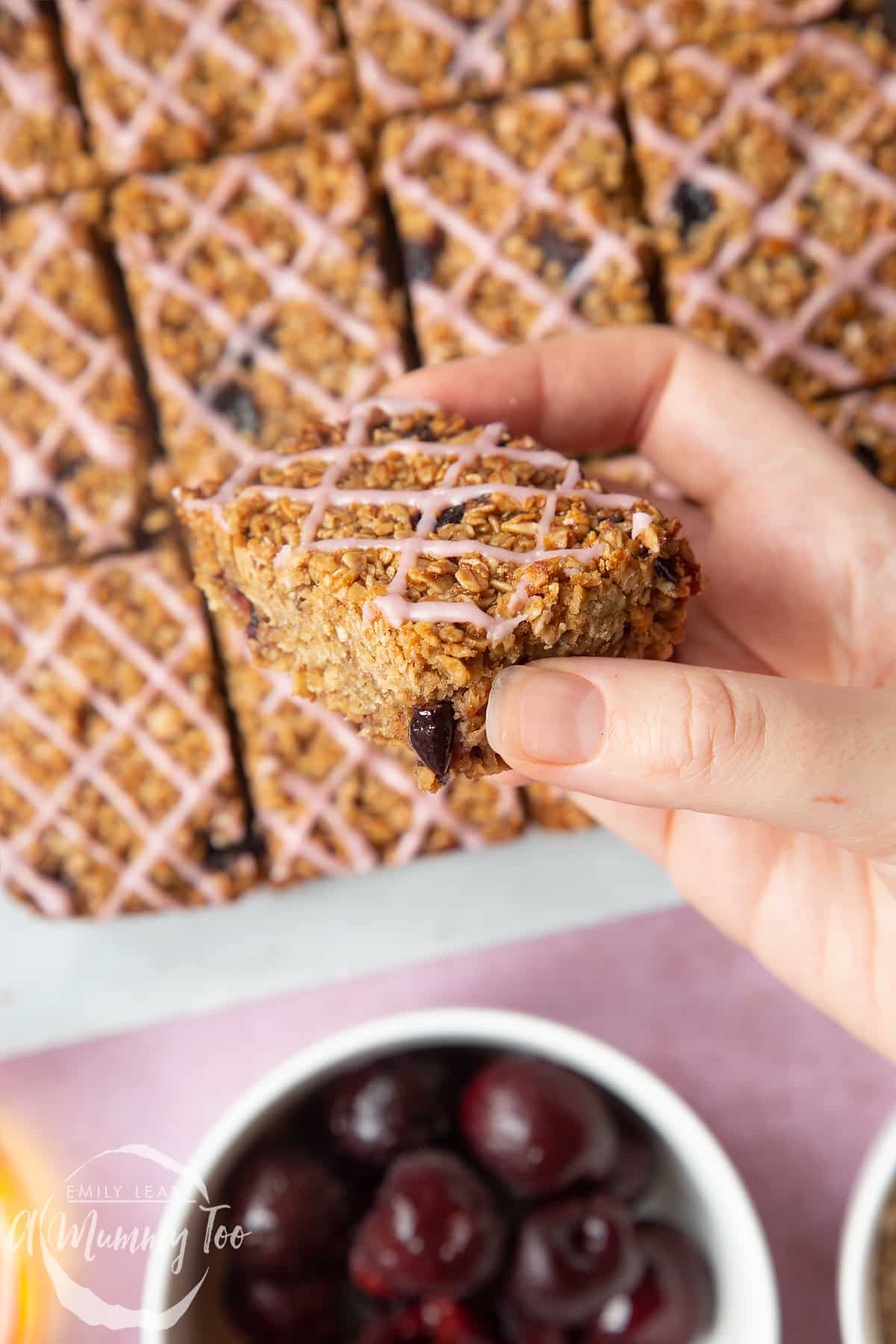 Overhead shot of a hand holding a cherry flapjack topped with pink icing served on a white plate with a mummy too logo in the lower-left corner