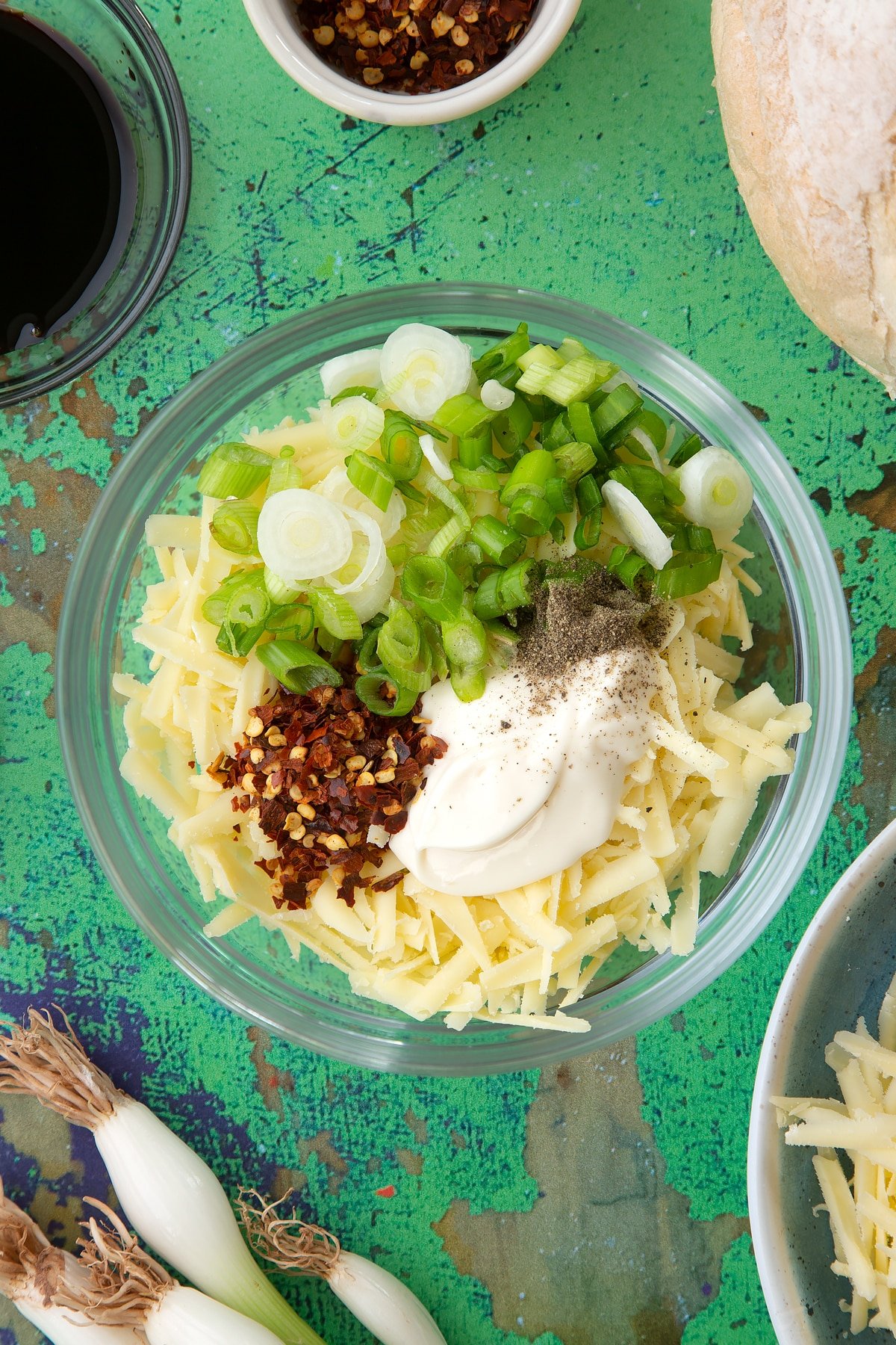 Cheddar, chilli flakes, spring onions, soy sauce, mayo and a pinch of pepper in a mixing bowl. Ingredients to make chilli cheese toast surround the bowl.