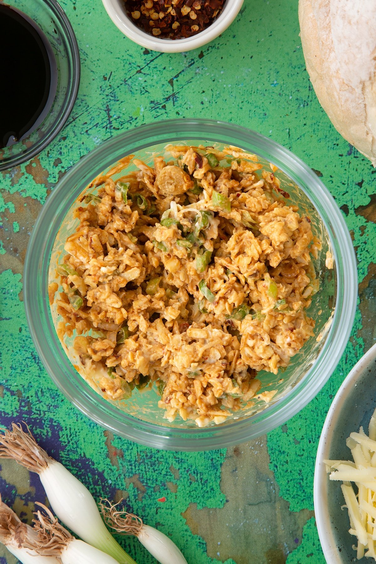 Cheddar, chilli flakes, spring onions, soy sauce, mayo and a pinch of pepper mixed together in a mixing bowl. Ingredients to make chilli cheese toast surround the bowl.