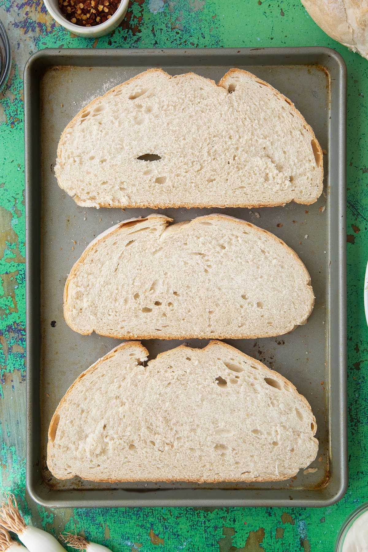 Three large slices of bread on a baking tray. Ingredients to make chilli cheese toast surround the tray.