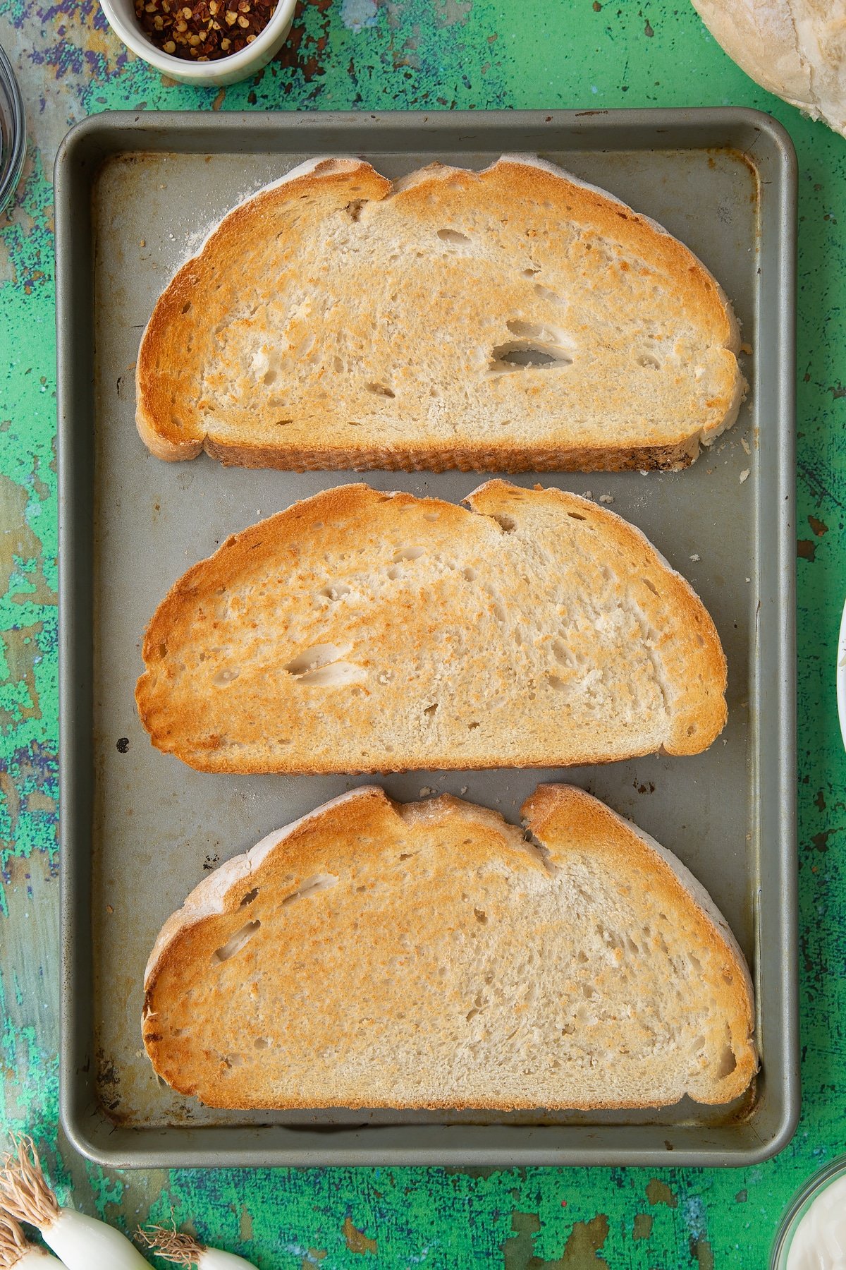 Three large slices of toast on a baking tray. Ingredients to make chilli cheese toast surround the tray.