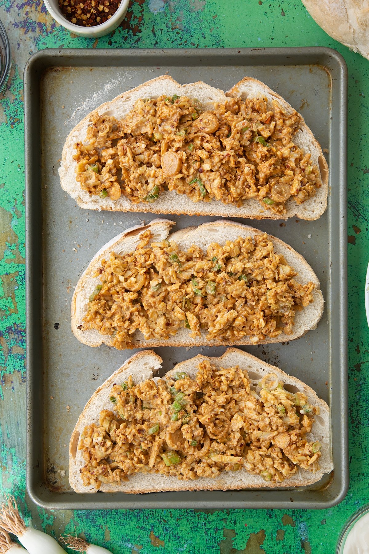 Three large slices of toast on a baking tray, spread with a mix of cheese, mayo and chilli. Ingredients to make chilli cheese toast surround the tray.