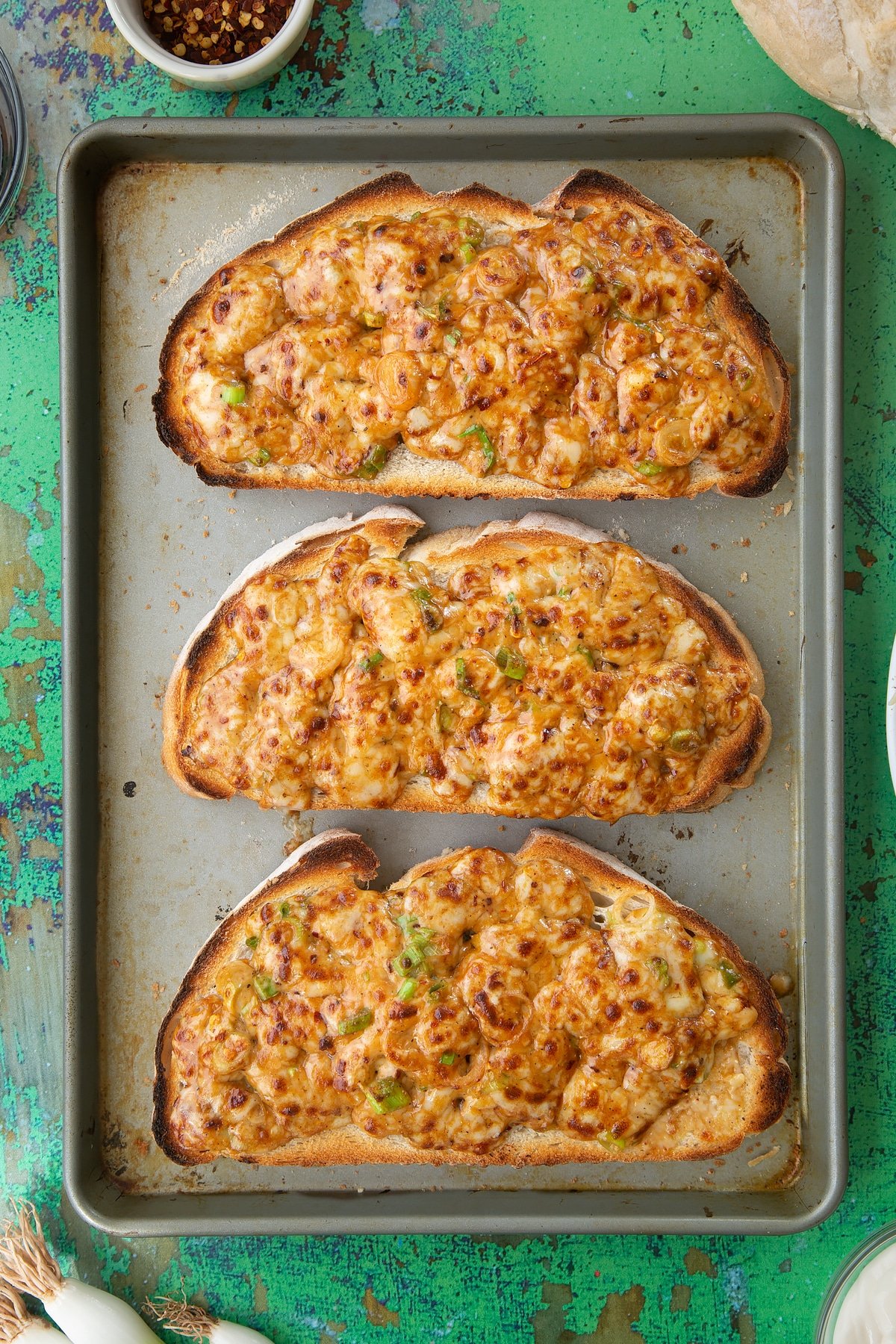 Three large slices of chilli cheese toast on a baking tray. Ingredients to make chilli cheese toast surround the tray.