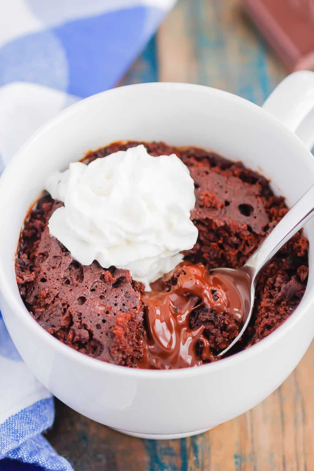 Close up of chocolate lava mug cake. The cake is topped with a small swirl of cream and a spoon delves in to reveal the lava centre.