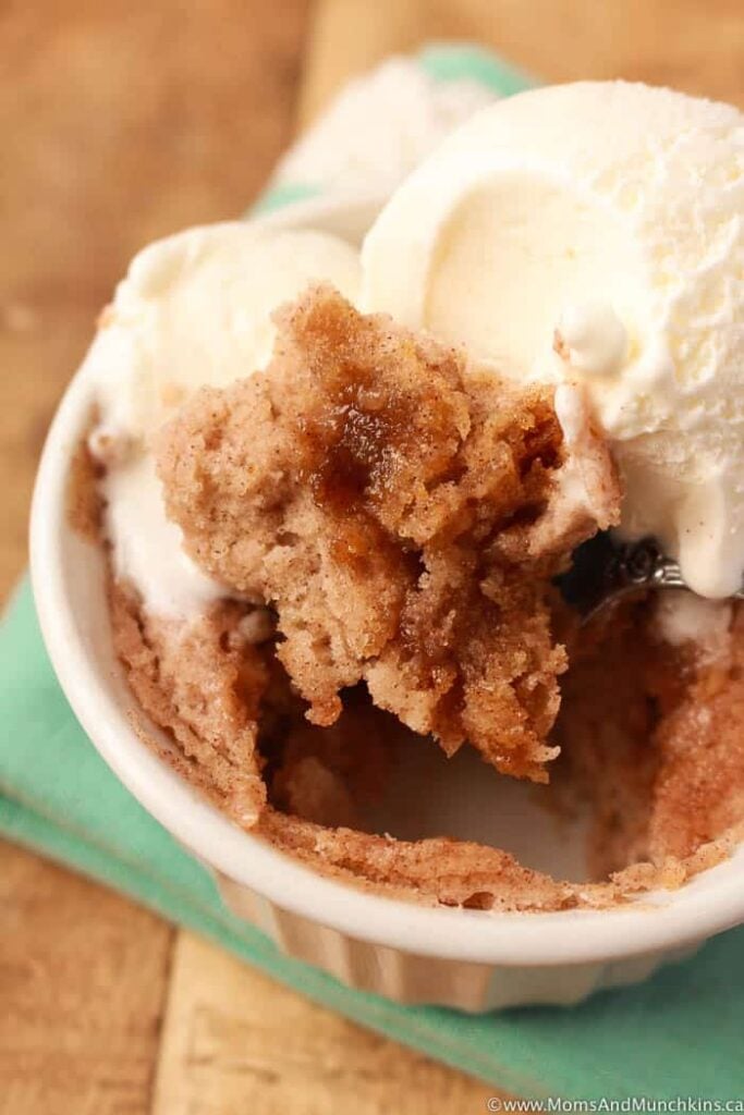 Cinnamon roll mug cake topped with vanilla ice cream. A spoon reveals the gooey cake below.