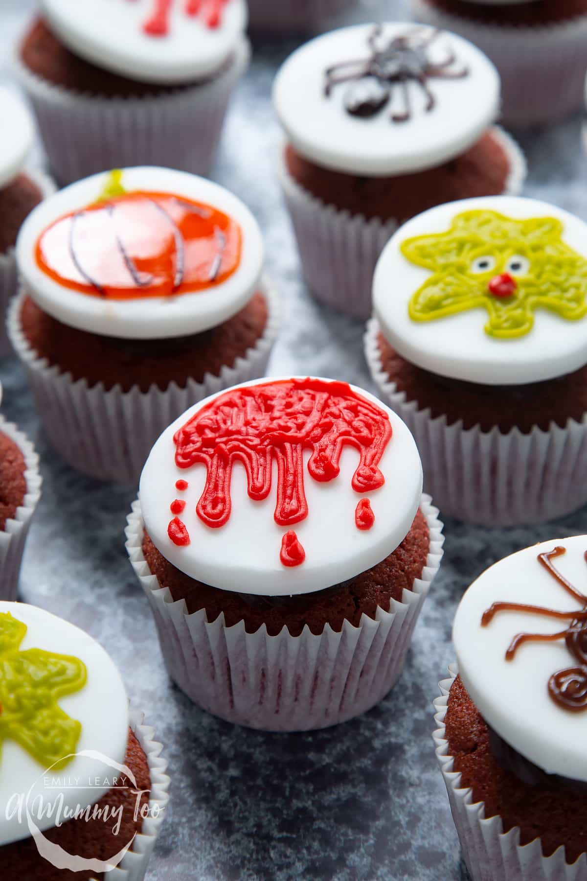 Dairy free Halloween cupcakes: red velvet cupcakes topped with chocolate frosting and white fondant discs decorated with icing pens. The cupcake in the foreground has a dripping blood design.