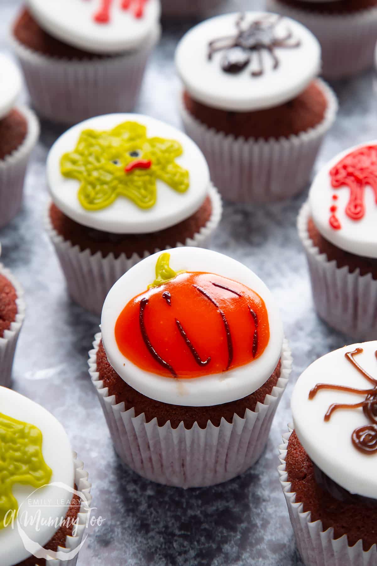 Dairy free Halloween cupcakes: red velvet cupcakes topped with chocolate frosting and white fondant discs decorated with icing pens. The cupcake in the foreground has a pumpkin design.