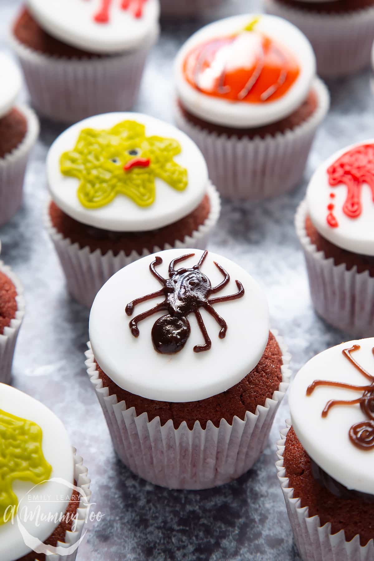 Dairy free Halloween cupcakes: red velvet cupcakes topped with chocolate frosting and white fondant discs decorated with icing pens. The cupcake in the foreground has a spider design.