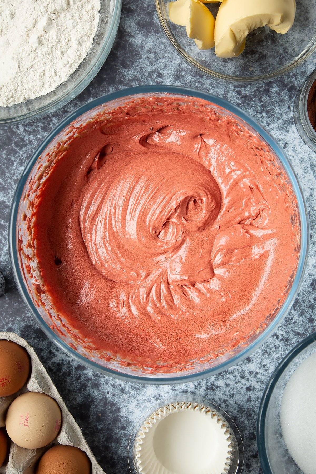 Dairy free red velvet cupcake batter and red food colouring in a large mixing bowl. Ingredients to make dairy free Halloween cupcakes surround the bowl.