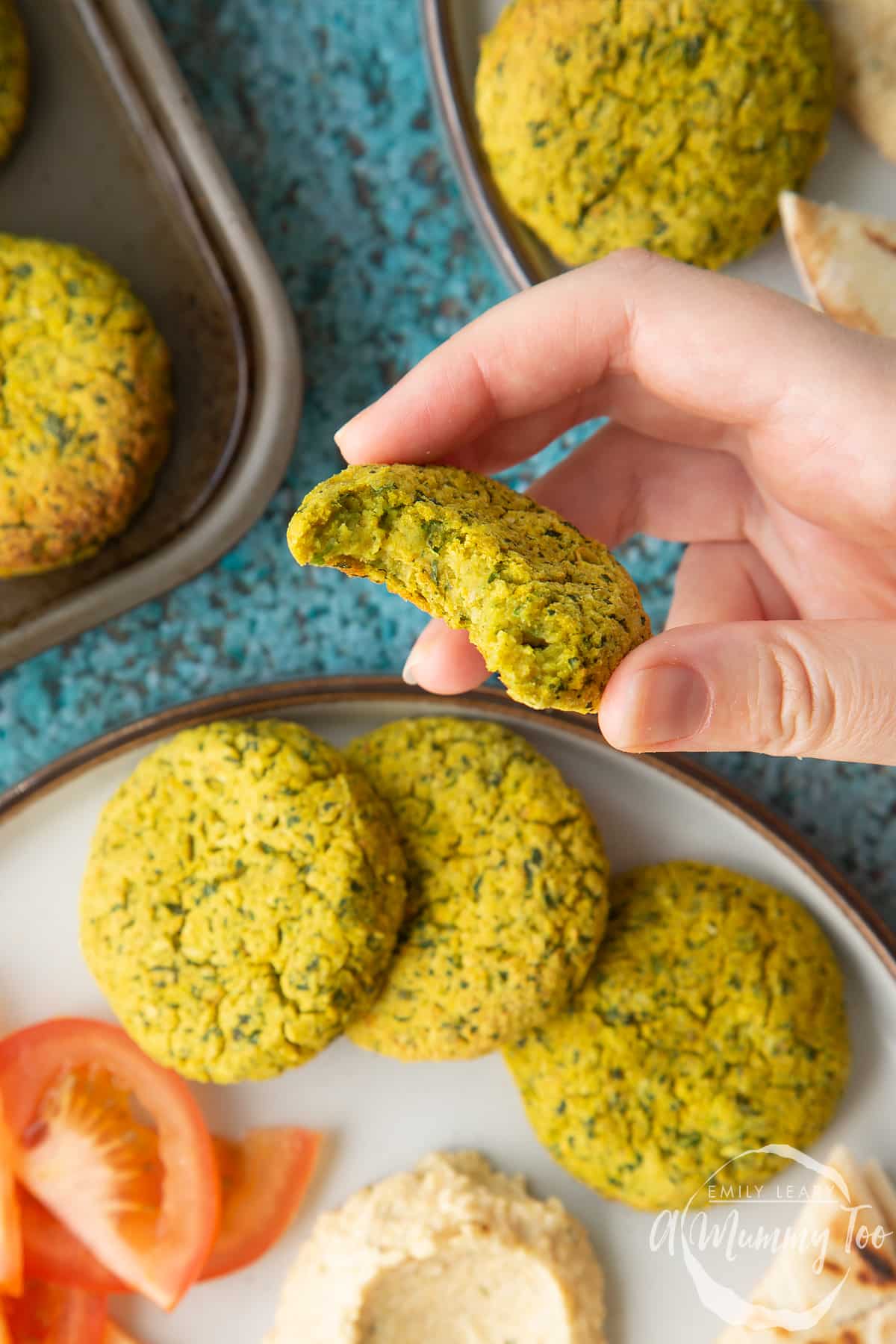 Hand holds a falafel with a bite out of it. In the background, gluten-free falafel on a plate with tomatoes, cucumber, hummus and griddled flatbread. 