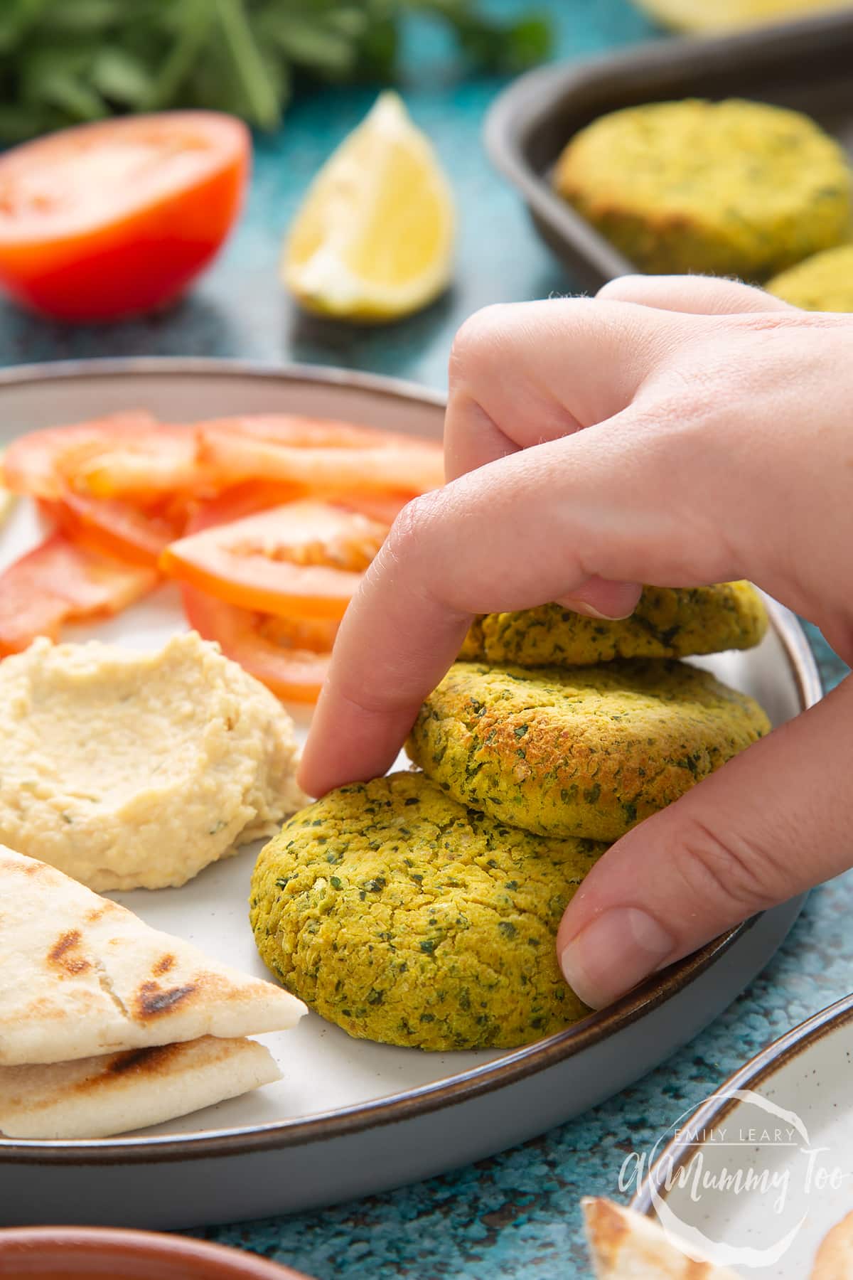 Gluten-free falafel arranged on a plate with tomatoes, cucumber, hummus and griddled flatbread. A hand reaches to take one.