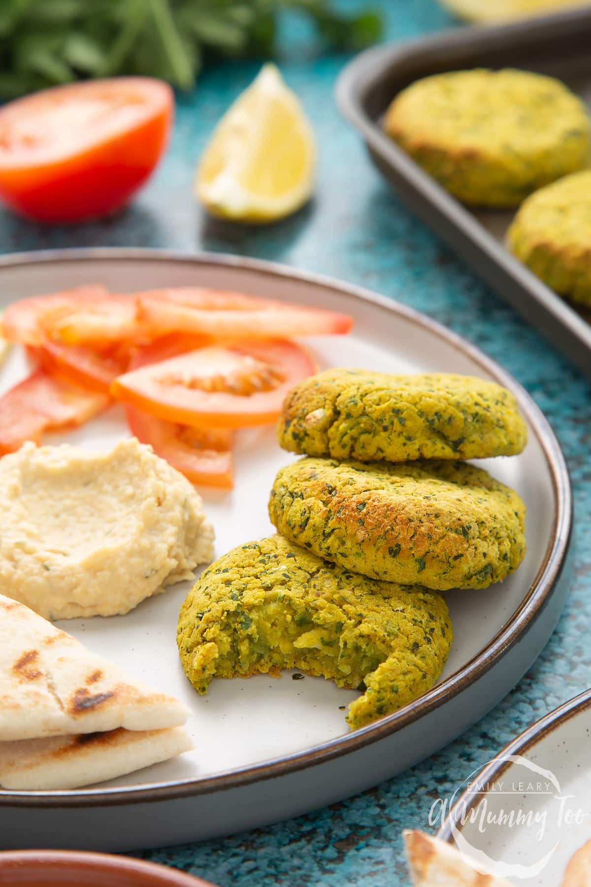 Gluten-free falafel arranged on a plate with tomatoes, cucumber, hummus and griddled flatbread. One of the falafel has a bite out of it.