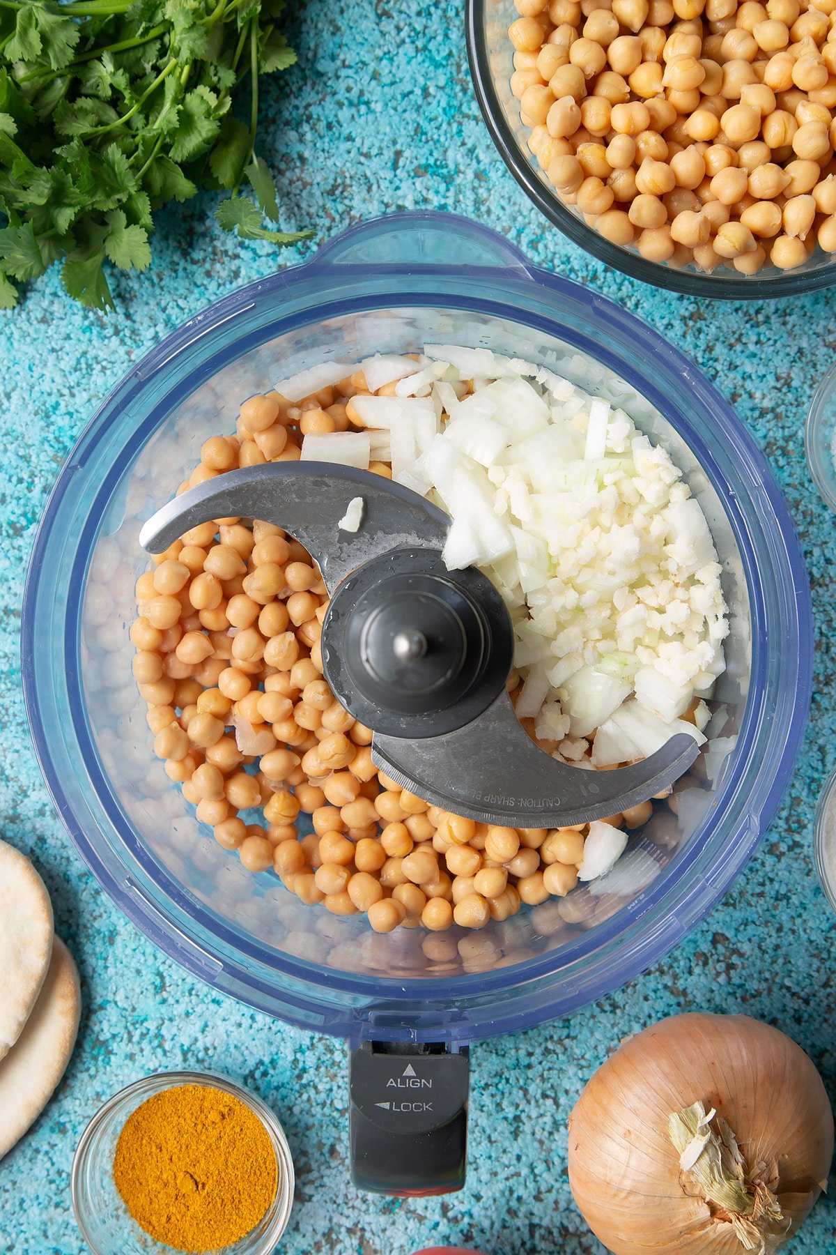 Chickpeas, onion and garlic in a food processor bowl. Ingredients to make gluten-free falafel surround the bowl.