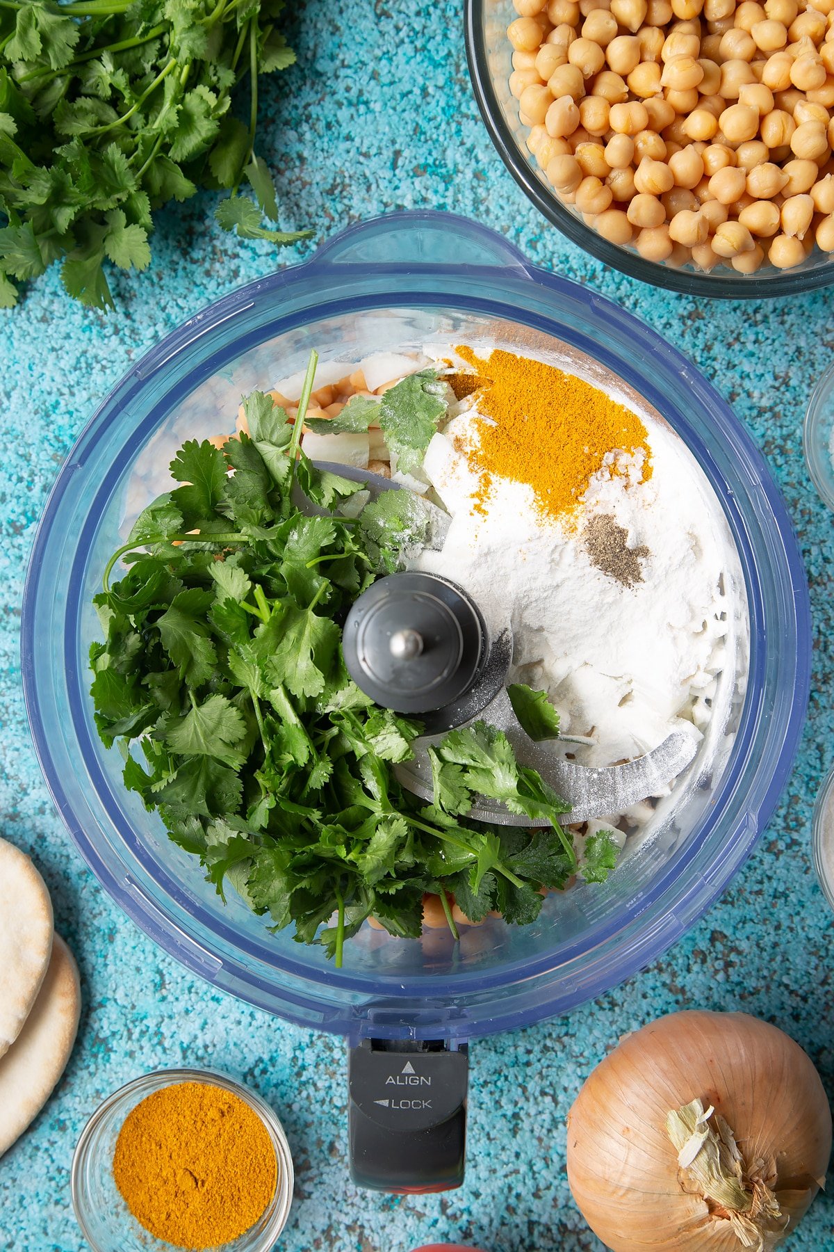 Chickpeas, onion, garlic, fresh coriander, gluten free flour, curry powder, salt and pepper in a food processor bowl. Ingredients to make gluten-free falafel surround the bowl.