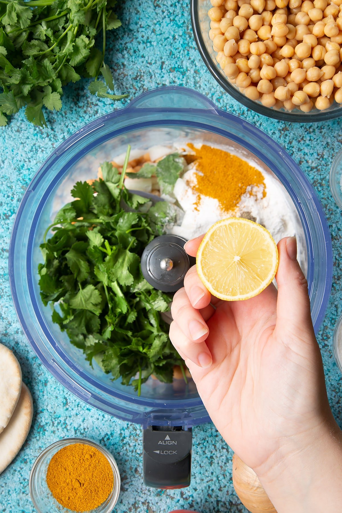 A hold holds a lemon half above chickpeas, onion, garlic, fresh coriander, gluten free flour, curry powder, salt and pepper in a food processor bowl. Ingredients to make gluten-free falafel surround the bowl.