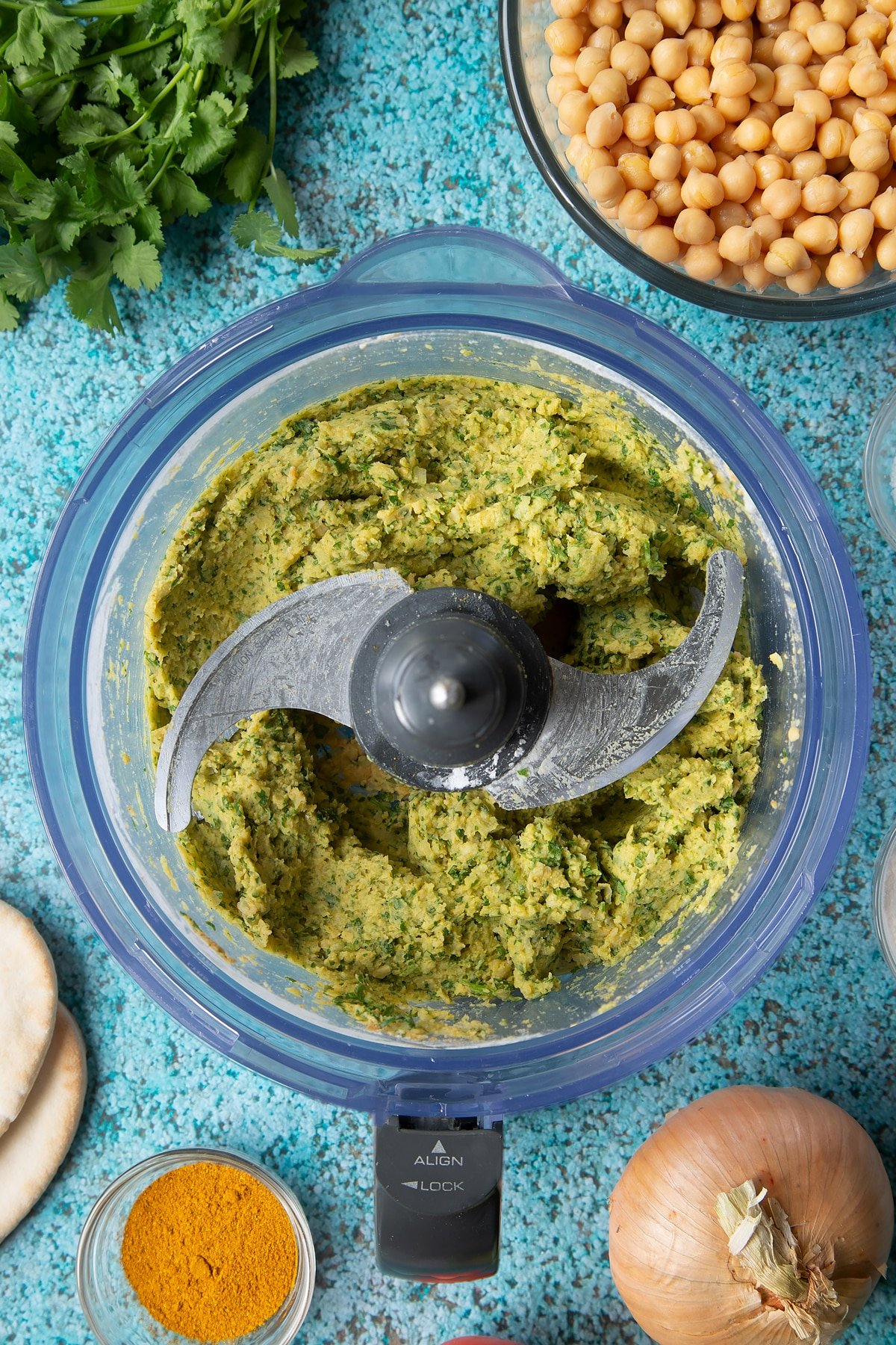 A paste made from chickpeas, coriander, onions, garlic and spices in a food processor bowl. Ingredients to make gluten-free falafel surround the bowl.