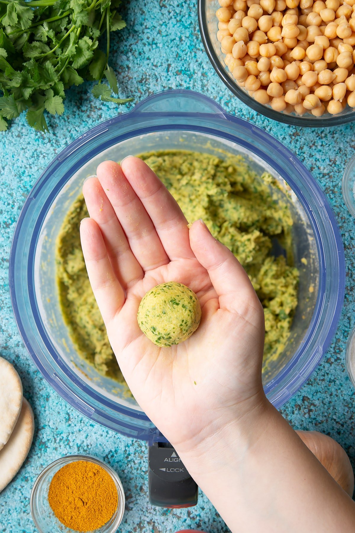 A hand holds a ball of falafel paste above a food processor bowl. Ingredients to make gluten-free falafel surround the bowl.