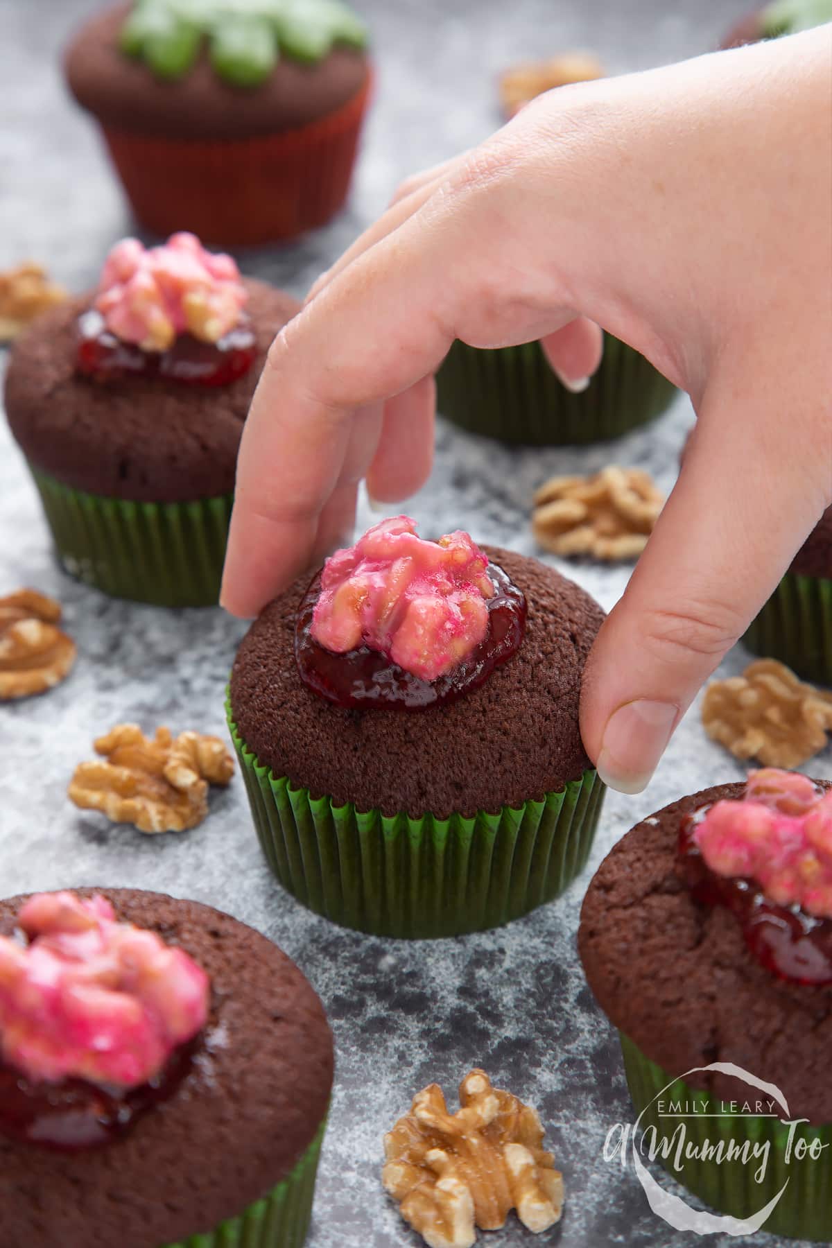 Gory Halloween cupcakes on a black background. The cupcakes are filled with jam and topped with pink candied walnuts that look like brains. A hand reaches for one.