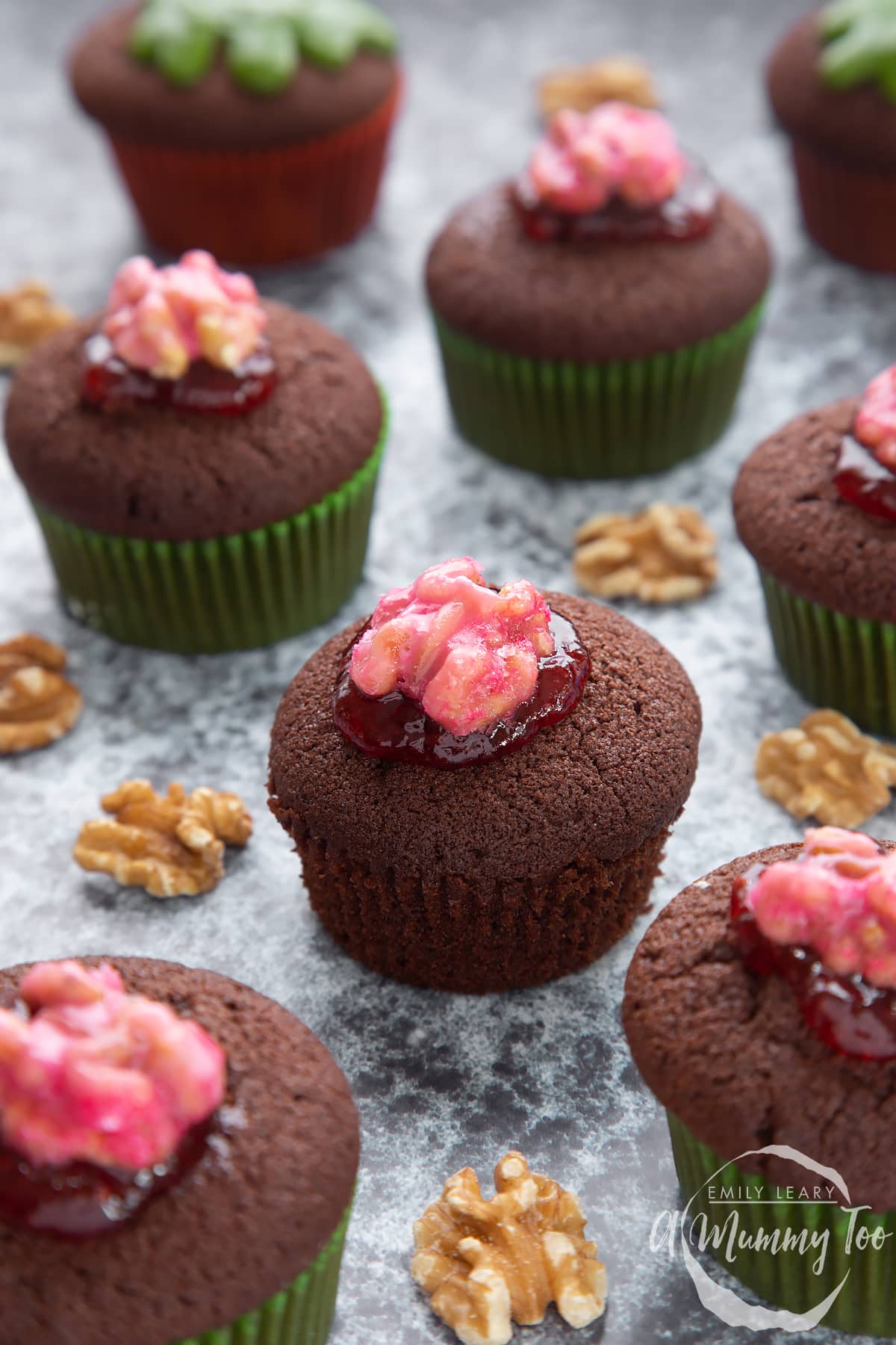 Gory Halloween cupcakes on a black background. The cupcakes are filled with jam and topped with pink candied walnuts that look like brains. One has been unwrapped.