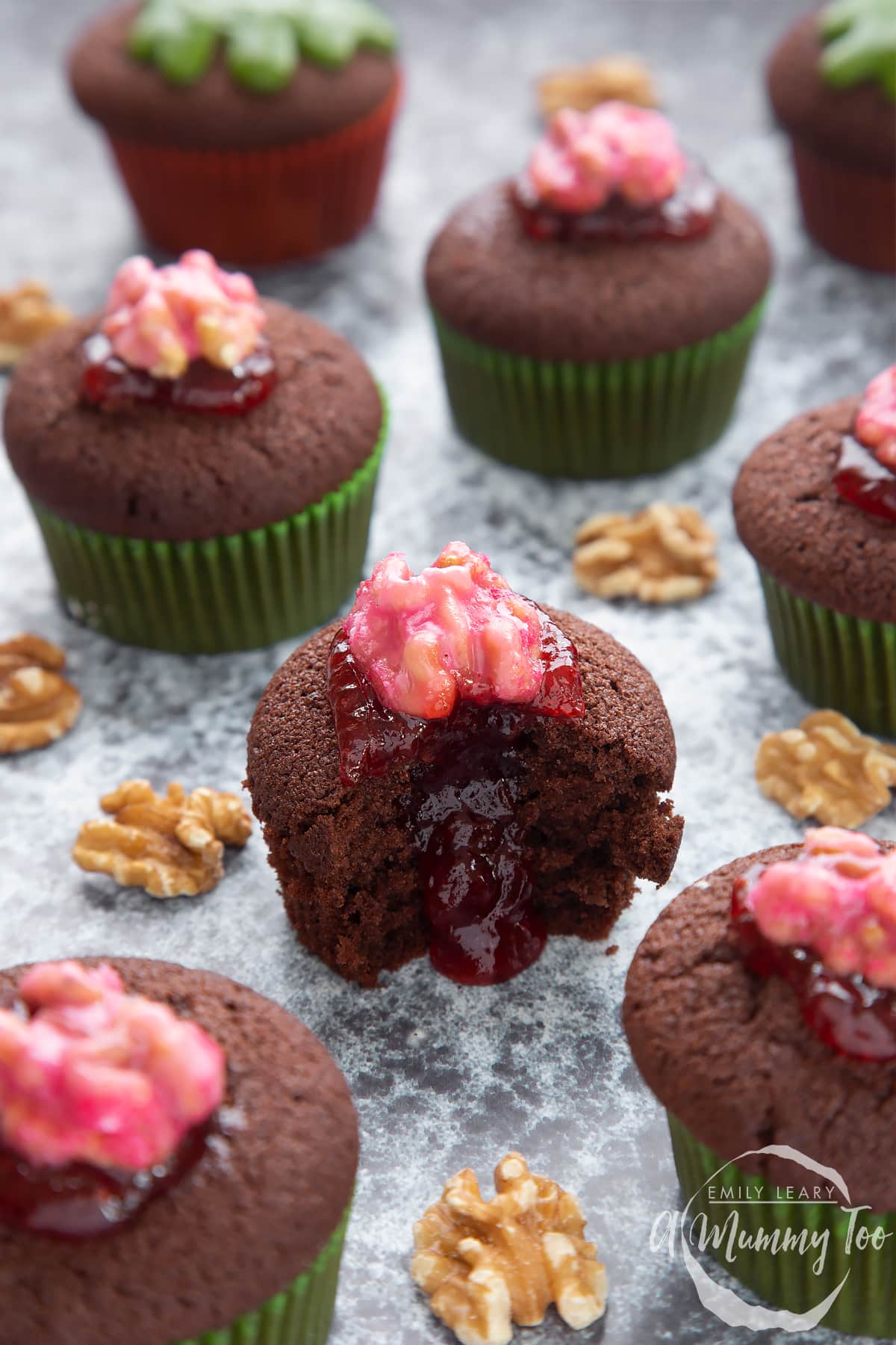 Gory Halloween cupcakes on a black background. The cupcakes are filled with jam and topped with pink candied walnuts that look like brains. The cupcake in the foreground has been bitten open revealing the jam core.