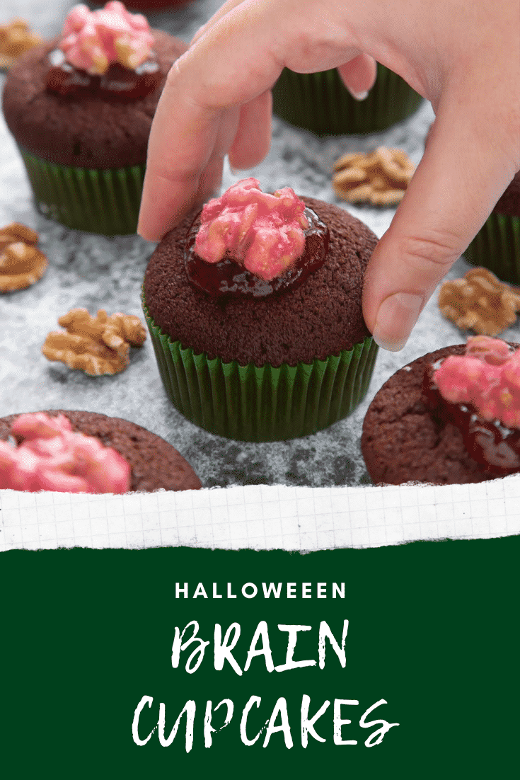 Gory Halloween cupcakes on a black background. The cupcakes are filled with jam and topped with pink candied walnuts that look like brains. A hand reaches to take one. Caption: Halloween brain cupcakes.