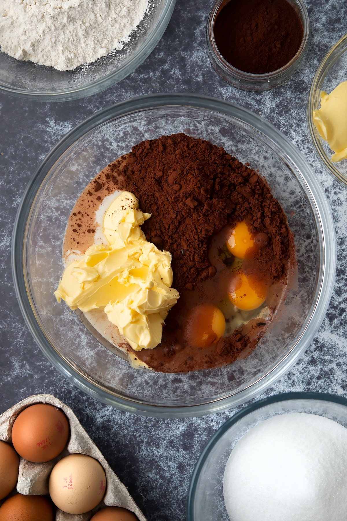 Margarine, oil, caster sugar, milk, cocoa and eggs in a large mixing bowl. Ingredients to make gory Halloween cupcakes surround the bowl.